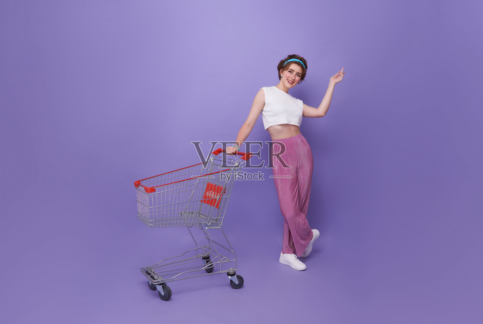在紫色的背景下，购物中心里，一个带着购物车的年轻快乐女人把食指放在一边。照片摄影图片