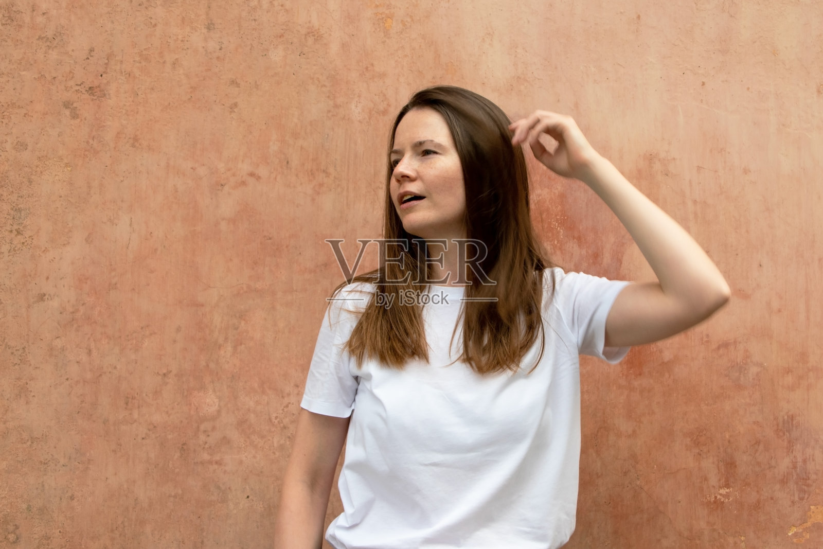 一个漂亮的年轻女人的肖像，棕色的长发，穿着白色的t恤，背景是水泥墙照片摄影图片