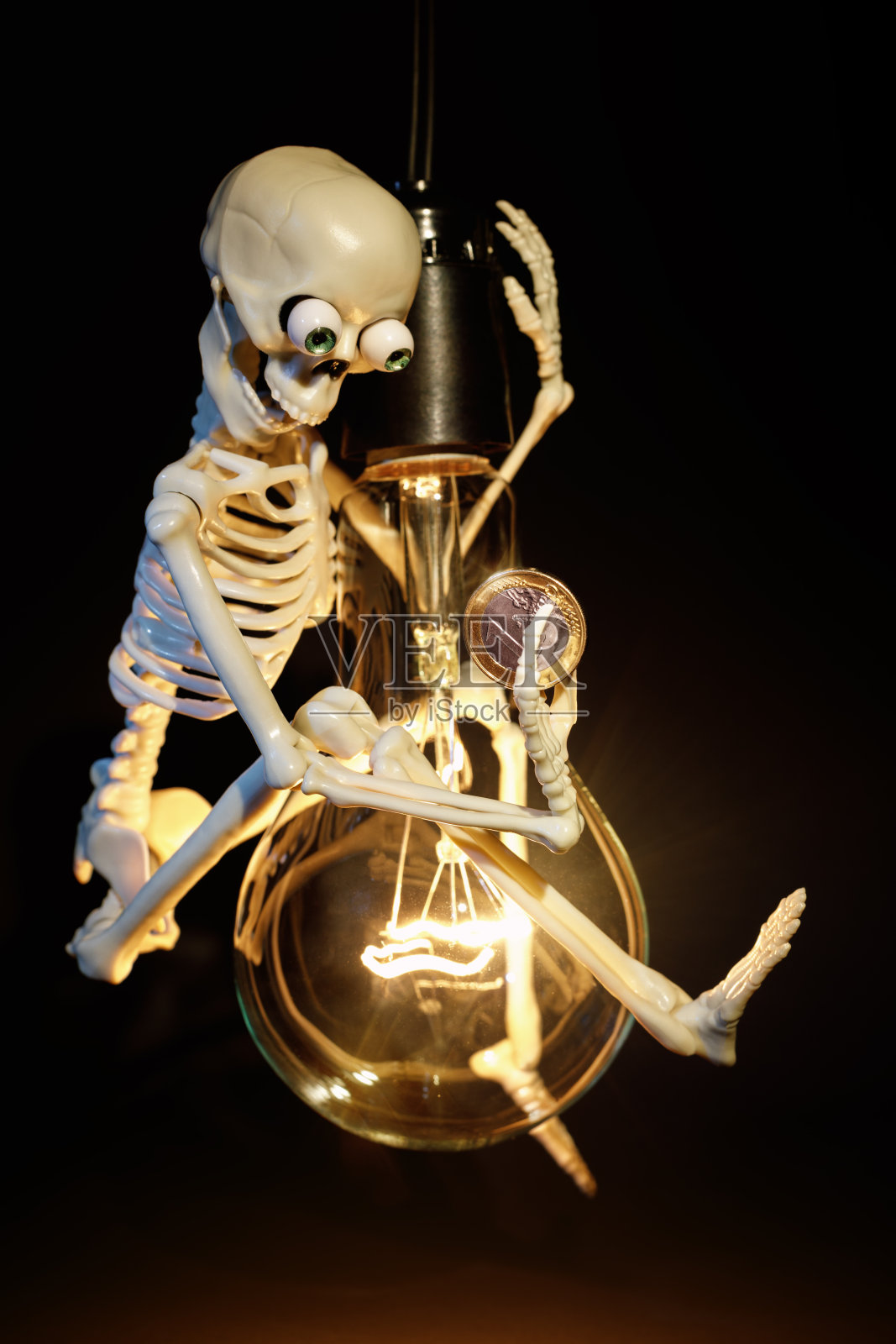一个有趣的玩具骷髅在一个黑色背景的大白炽灯泡上摇摆，他手里拿着一欧元用来付电费。图像有文字的空间照片摄影图片