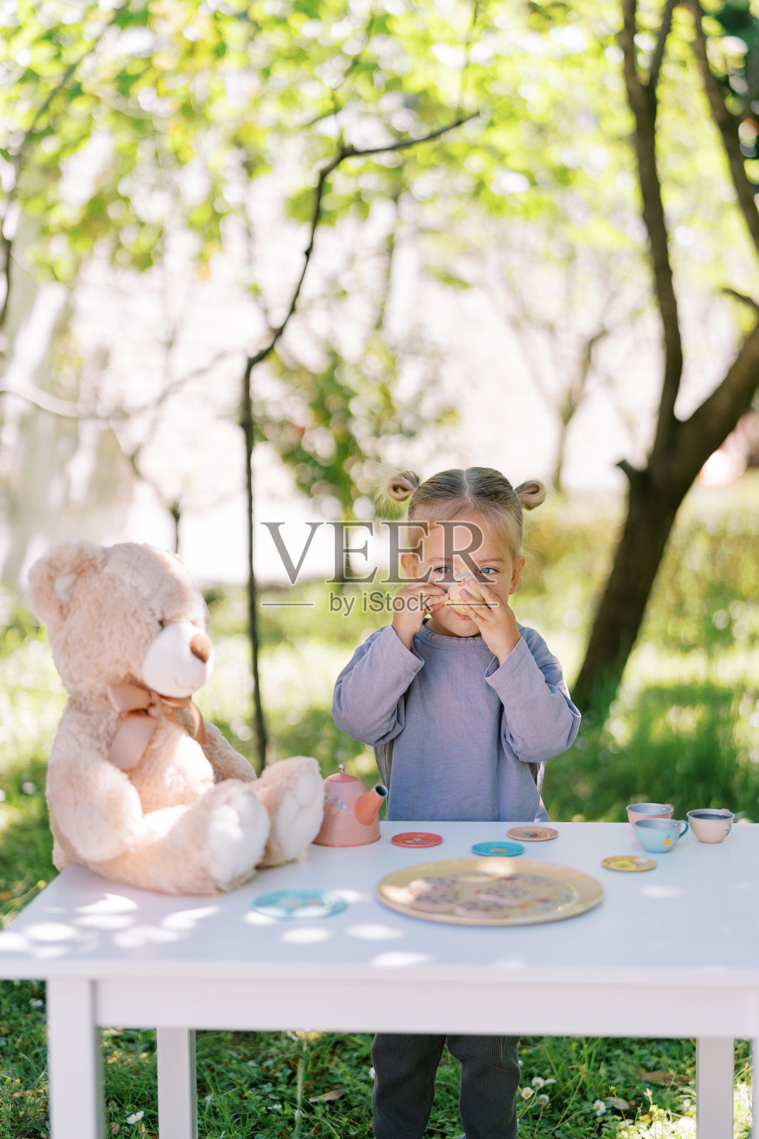茶话会上，小女孩用一个杯子喝茶，桌子上有一只泰迪熊照片摄影图片
