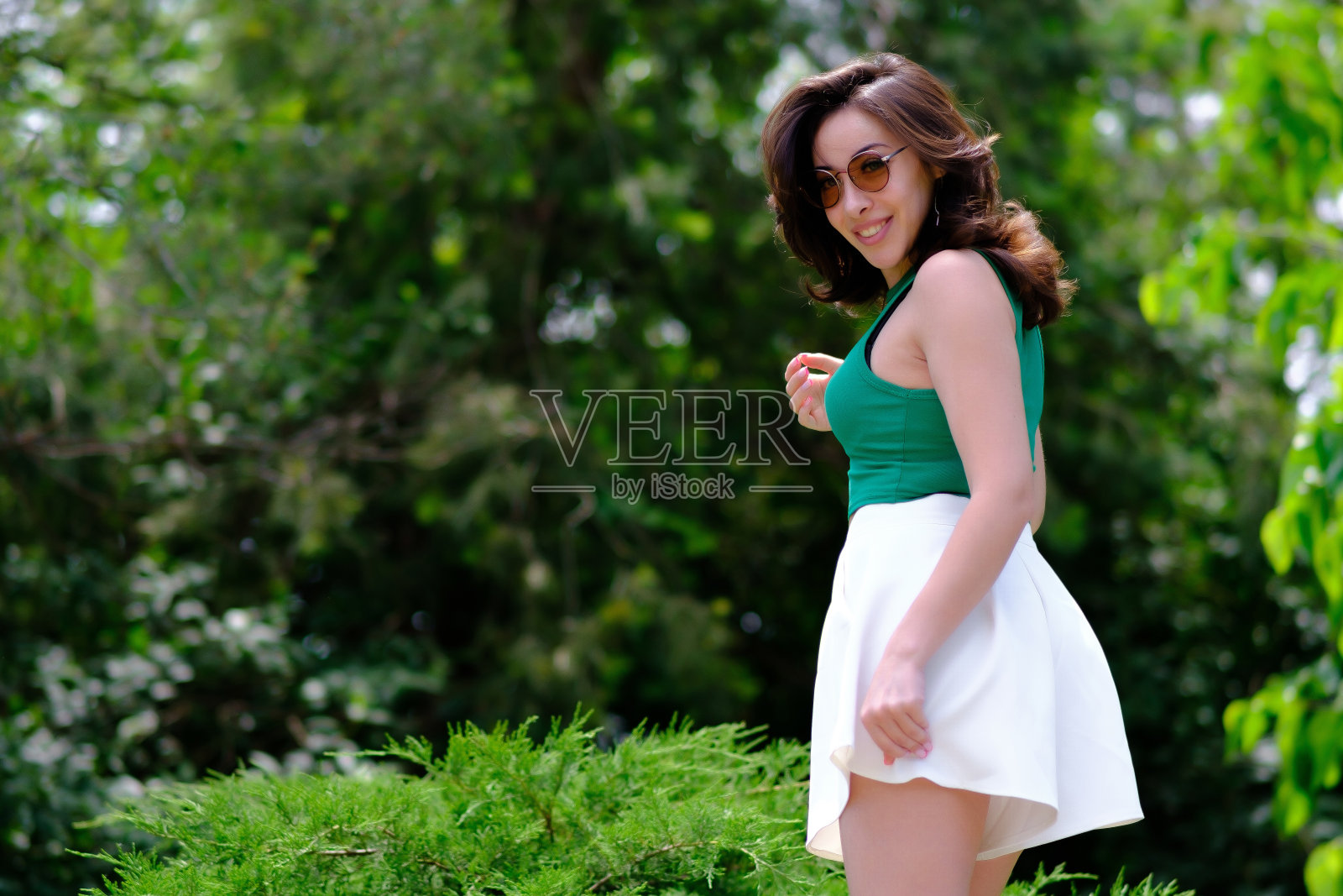 在一个阳光明媚的夏日，一个美丽的女孩穿着绿色上衣和白色短裤在城市公园里摆姿势照片摄影图片