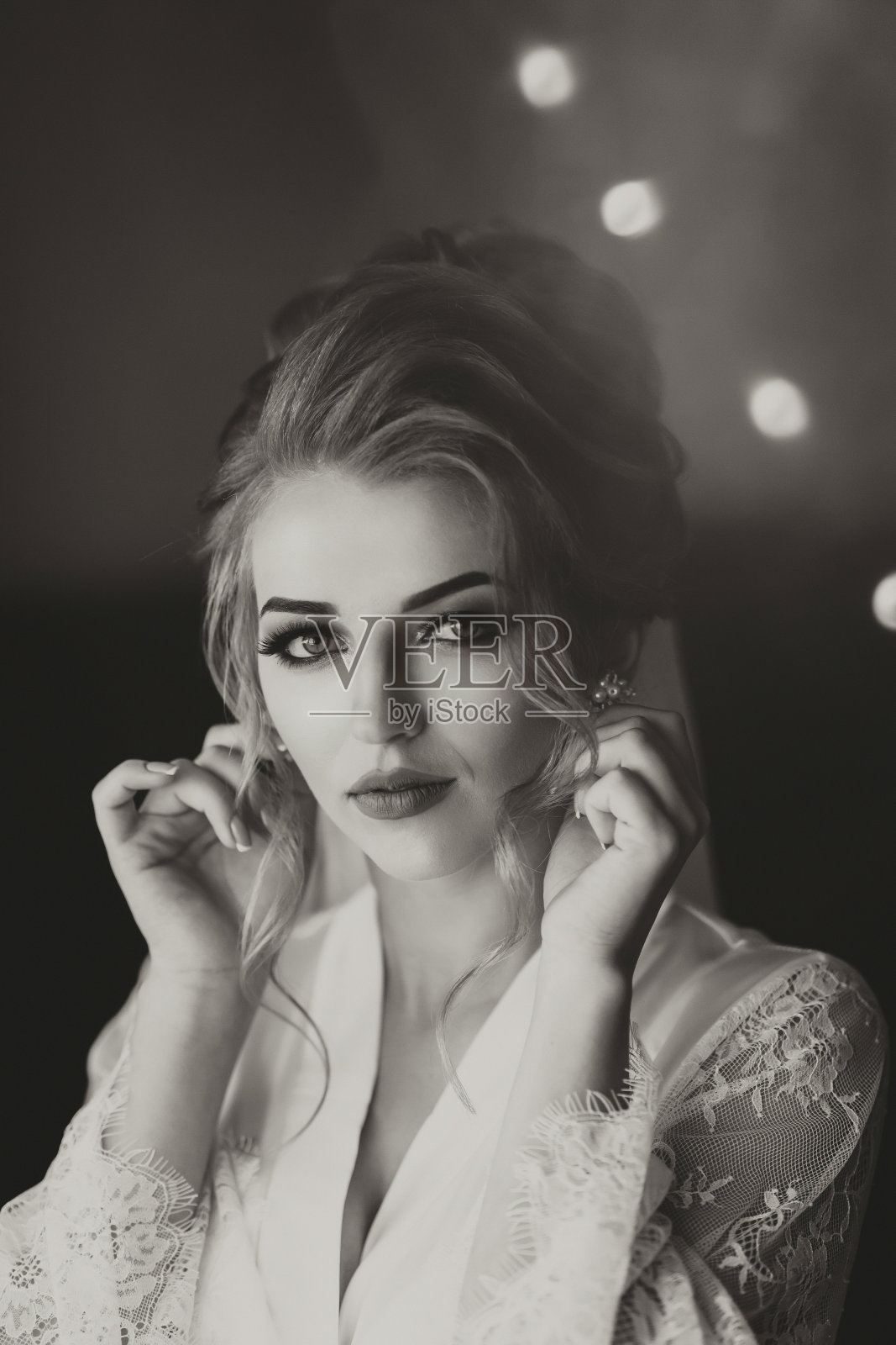 黑白照片。一个金发碧眼的新娘穿着一条长长的蕾丝衬裙，站在她的房间里，在镜子前摆姿势，戴上耳环。漂亮的发型和妆容，敞开胸围。婚礼的肖像。镜子里的倒影照片摄影图片