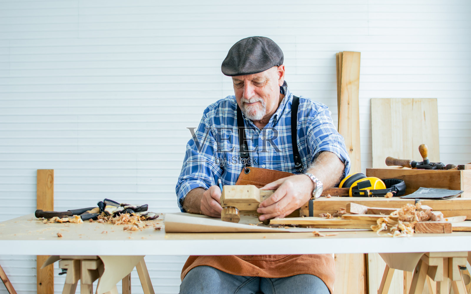 资深高加索快乐退休老木工或木匠微笑着，穿着格子衬衫，系着围裙，戴着复古帽，DIY木制迷你人偶玩具作为退休后的爱好装饰。照片摄影图片