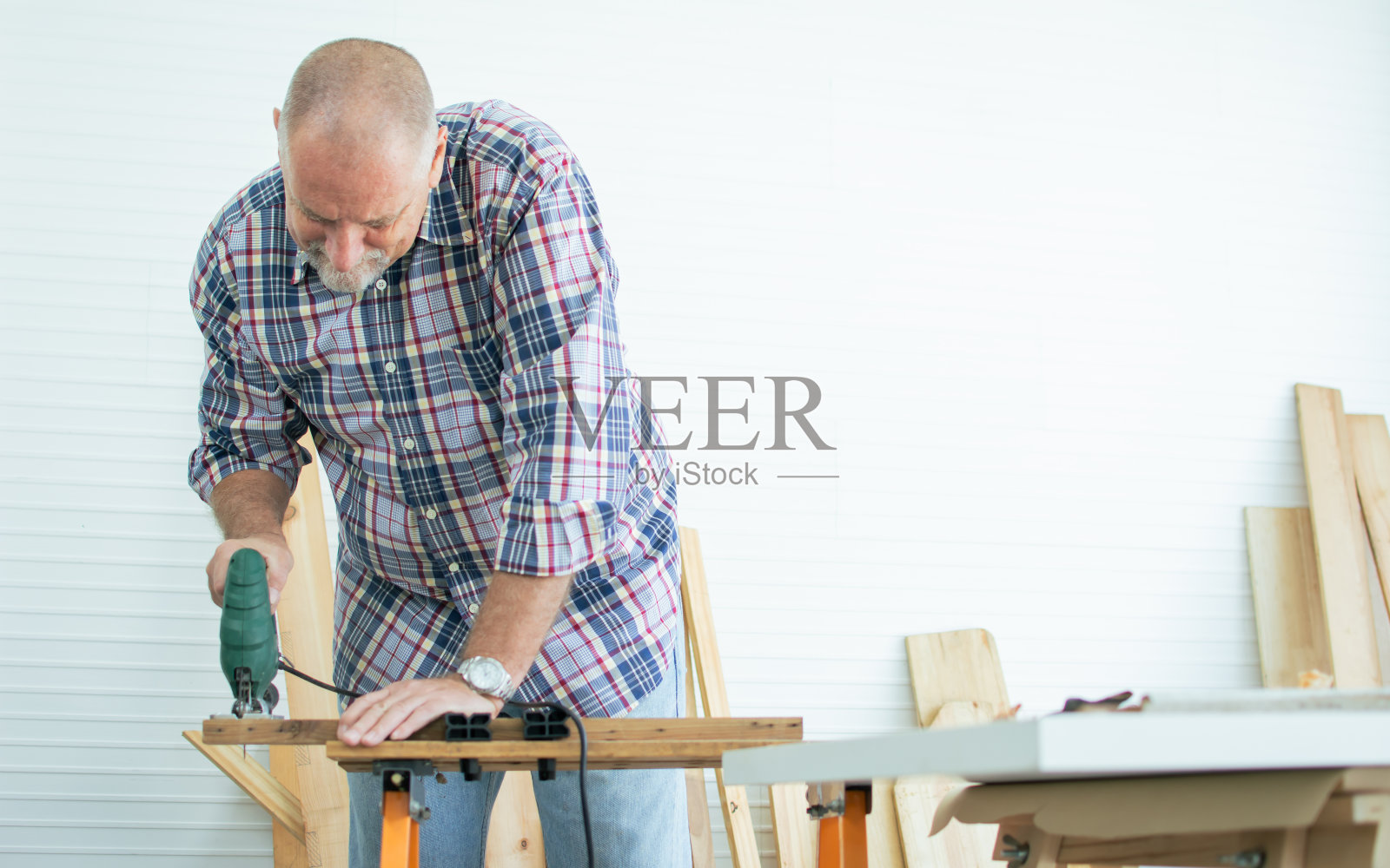 老白种人穿着格子衬衫，系着围裙，自己动手做木制家具，一个人站在家里，开心地用设备钻木头。退休与爱好概念。照片摄影图片