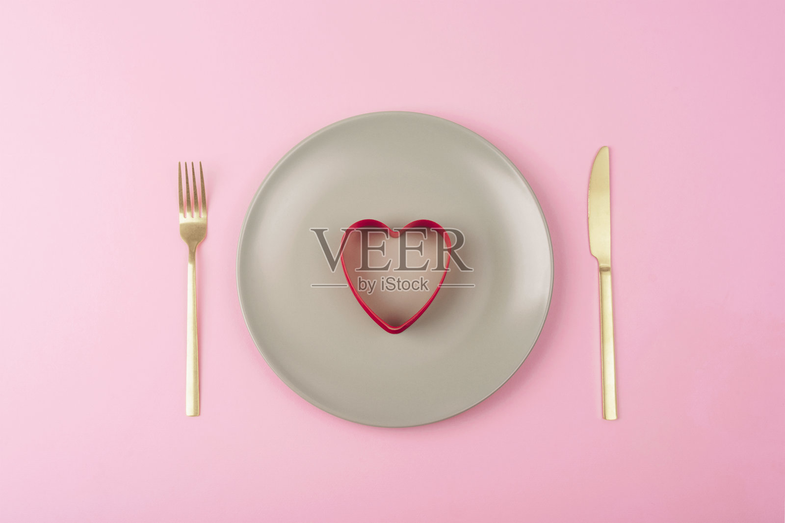 灰色的盘子上有心形的红色饼干切刀，粉红色的背景上有刀叉。情人节概念。俯视图，平铺照片摄影图片