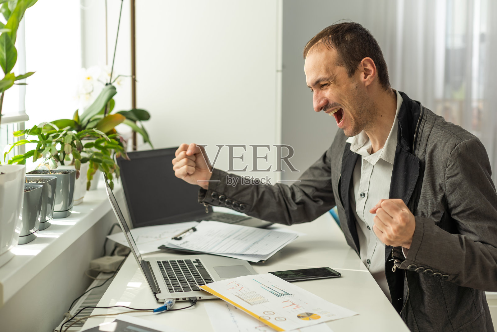 胜利的概念。一位成功、快乐、幸运、留着胡子的白人商人、公司首席执行官、经理，坐在办公室的办公桌前，用拳头打手势照片摄影图片