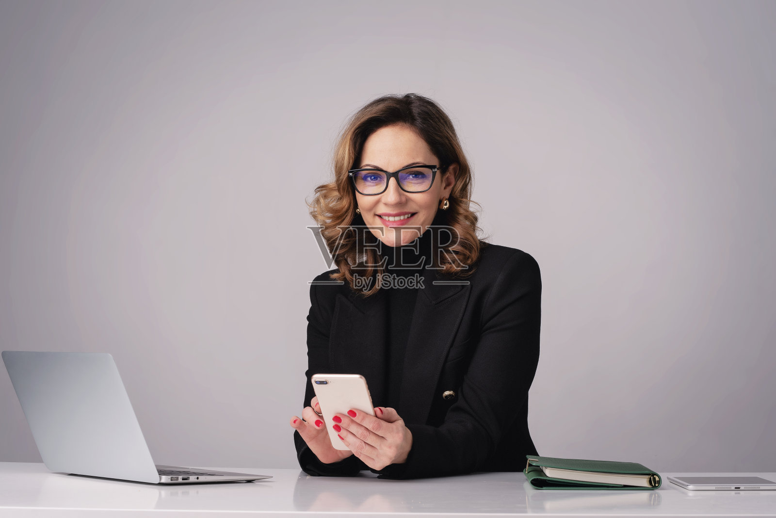 成熟的女性坐在办公桌前，在孤立的背景下使用笔记本电脑和智能手机照片摄影图片