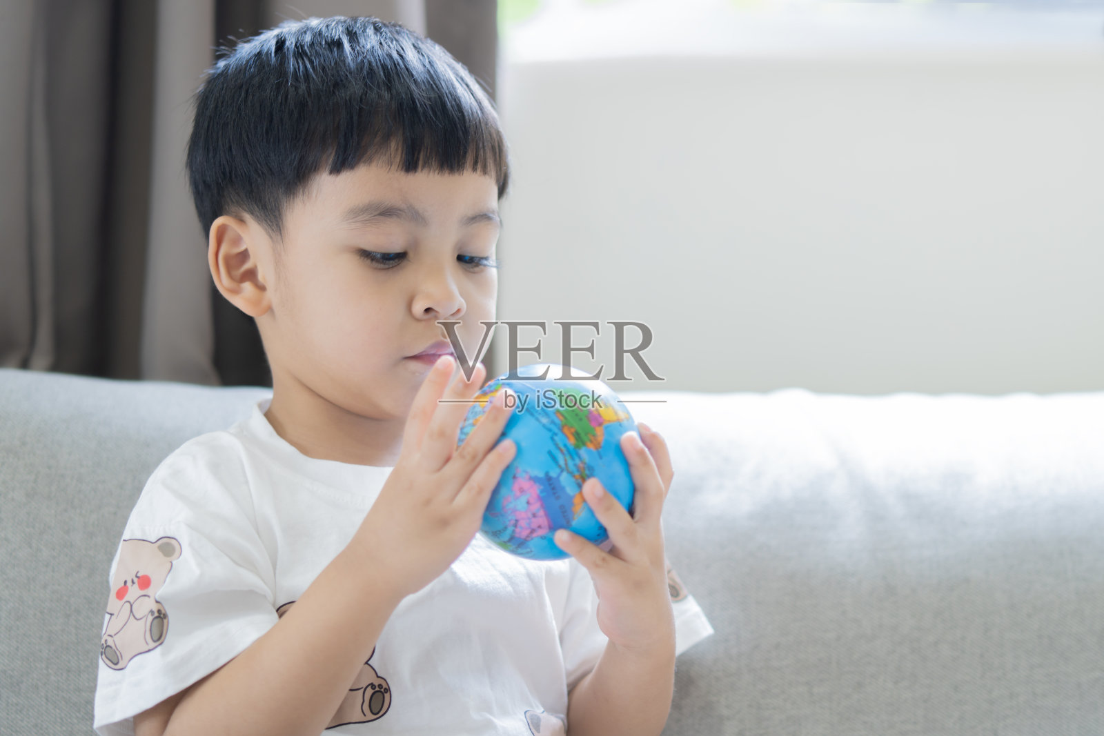 亚洲小男孩坐在沙发上玩地球球，真正的阳光从家里的窗户照进来。照片摄影图片