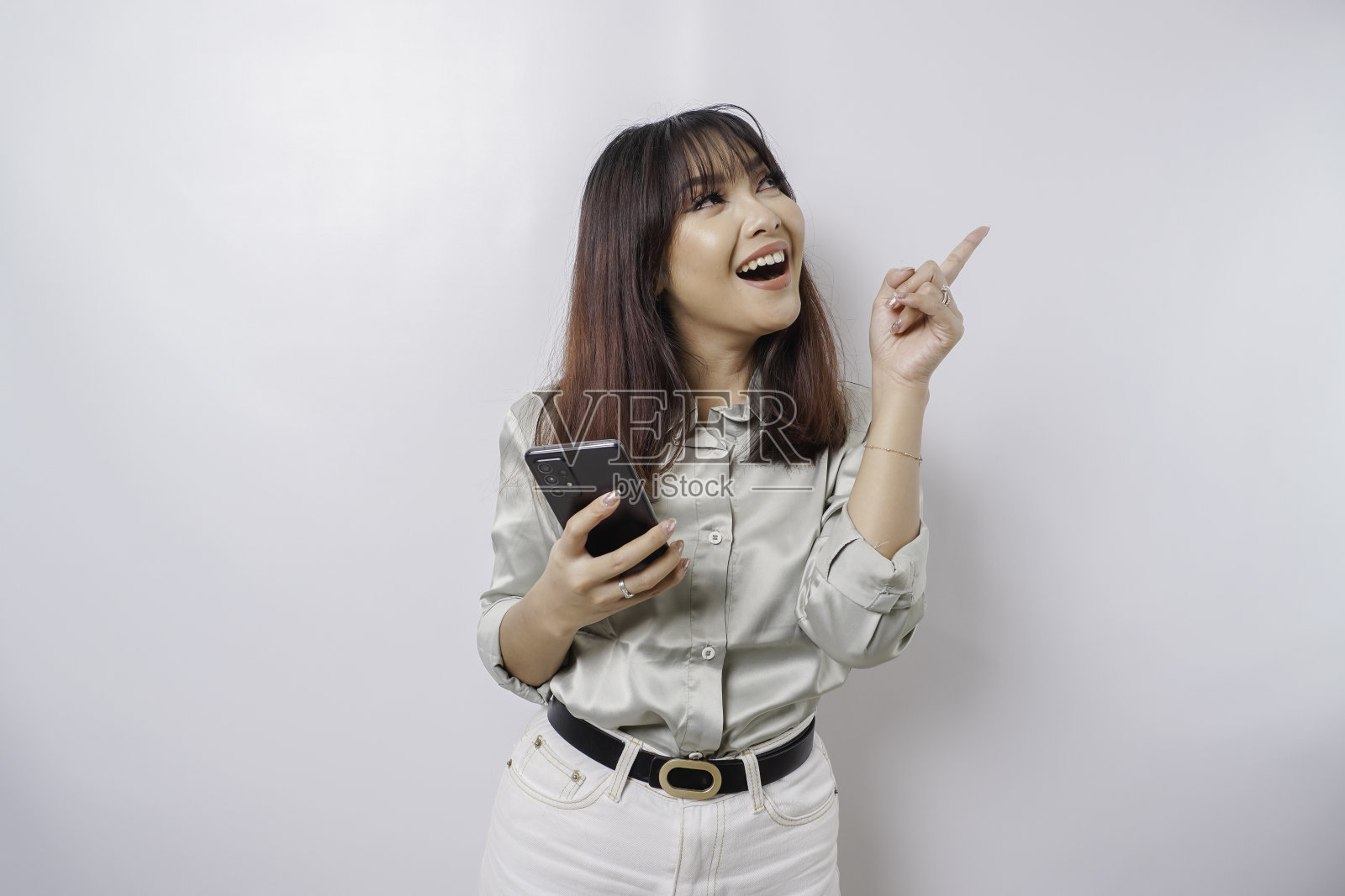 一名穿着鼠尾草绿色套装的亚洲女子兴奋地指着她身上的复制空间，手里拿着手机，被白色的背景隔开照片摄影图片