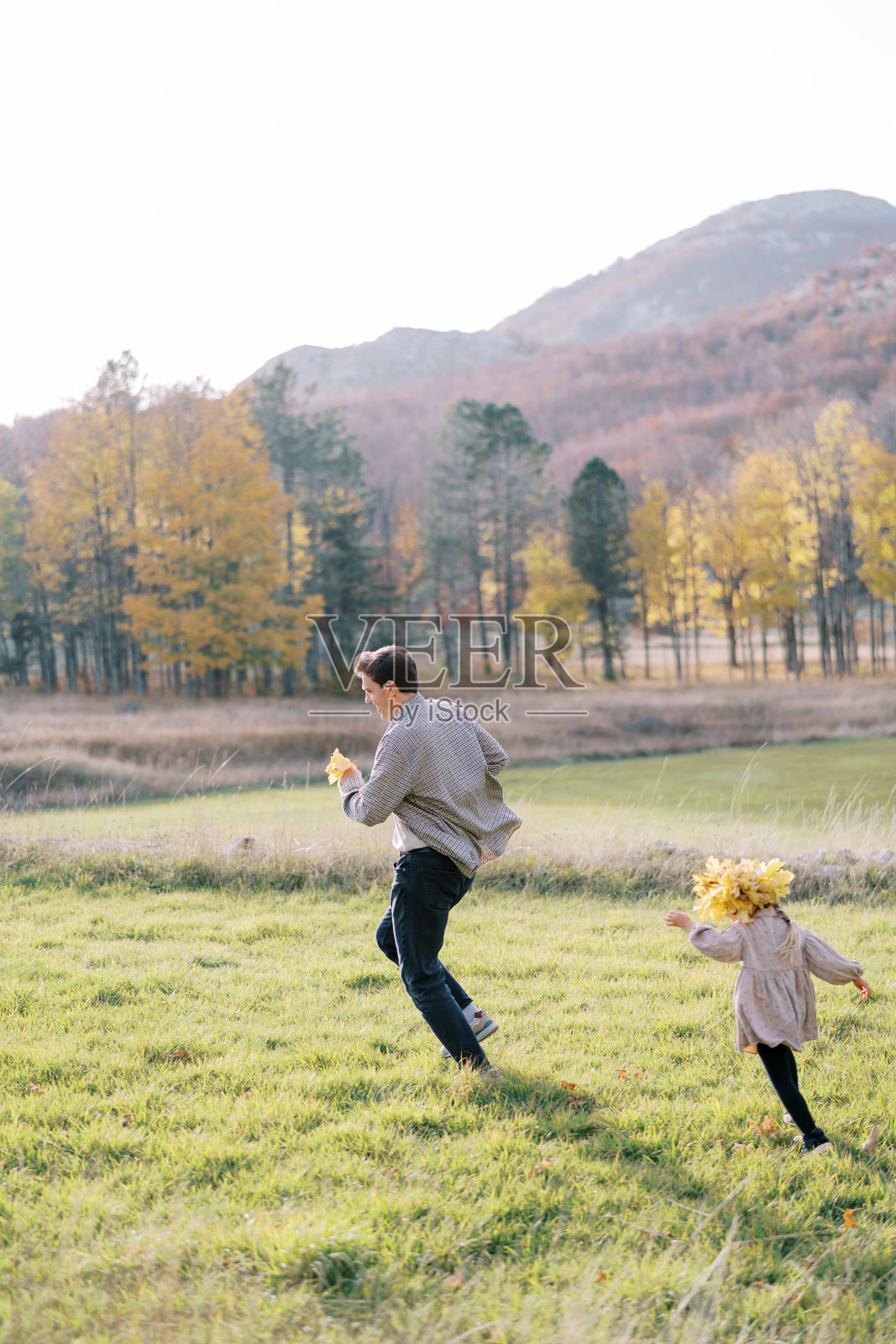 小女孩戴着秋叶花环，在阳光明媚的草坪上追着爸爸跑照片摄影图片