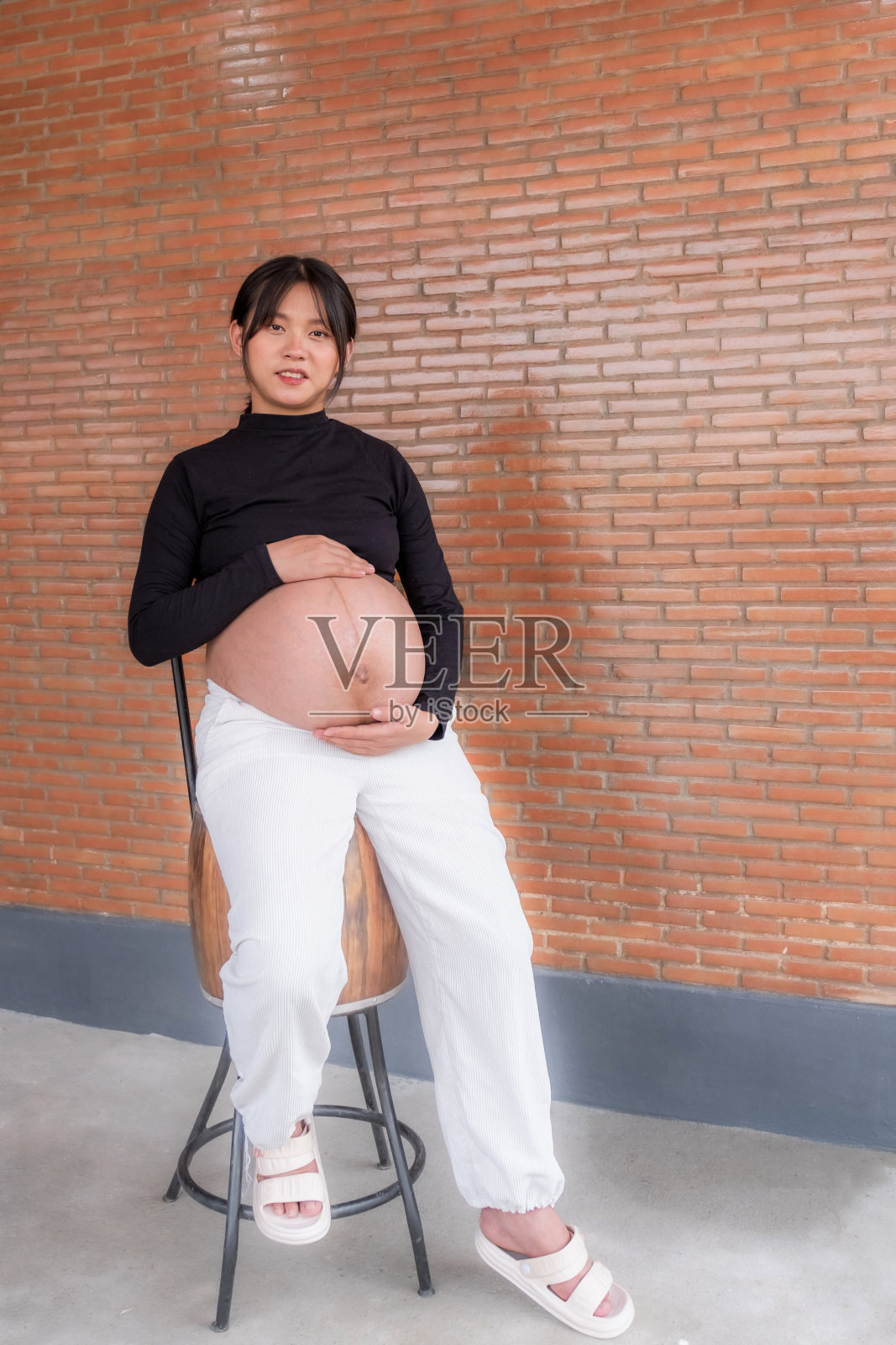 穿着裙子的孕妇妈妈在红砖背景上把手放在肚子上，用爱等待着你的宝宝的诞生照片摄影图片