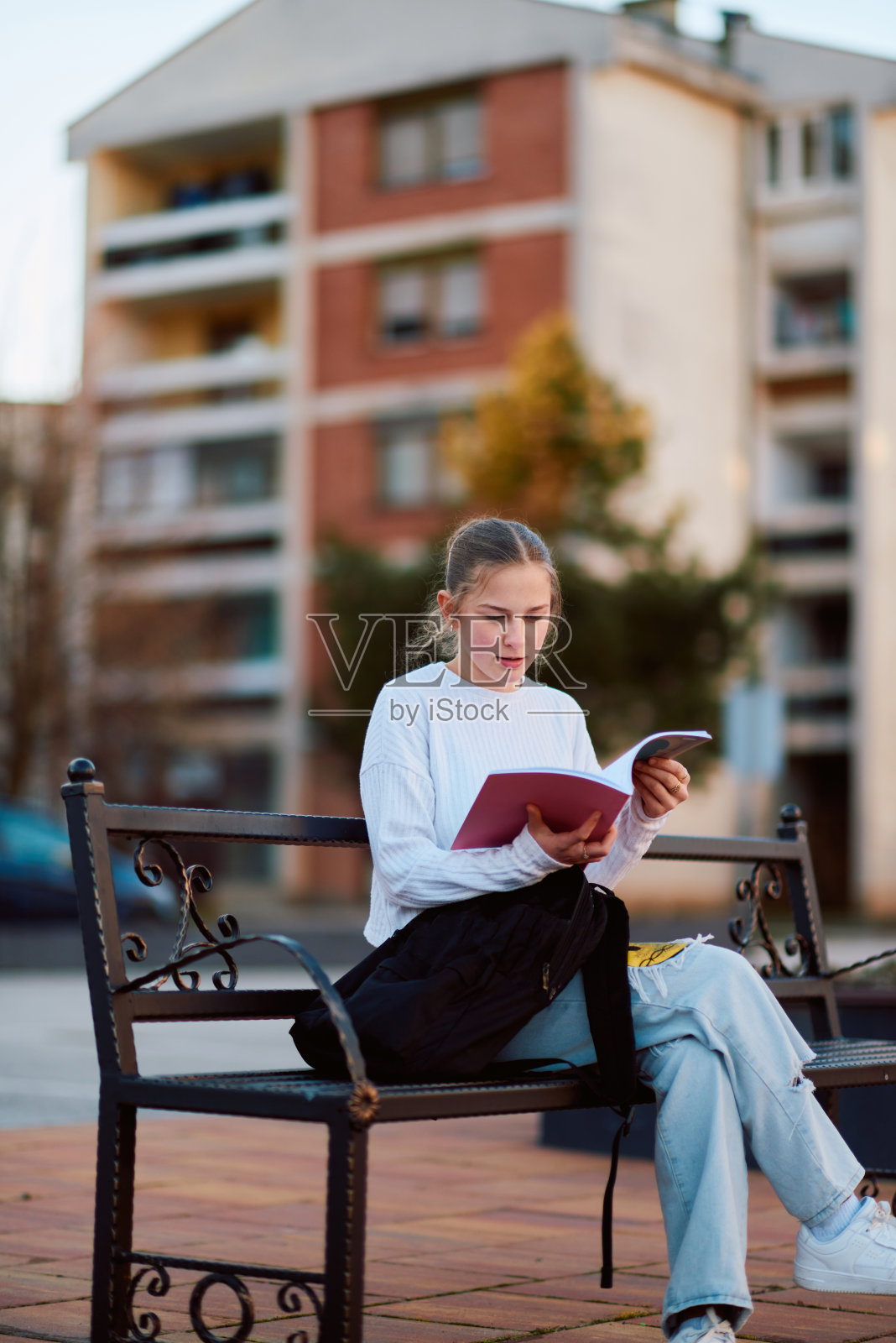 一位金发少女坐在公园的长椅上，从学校休息一下，沉浸在一本书中，旁边是她的书包照片摄影图片