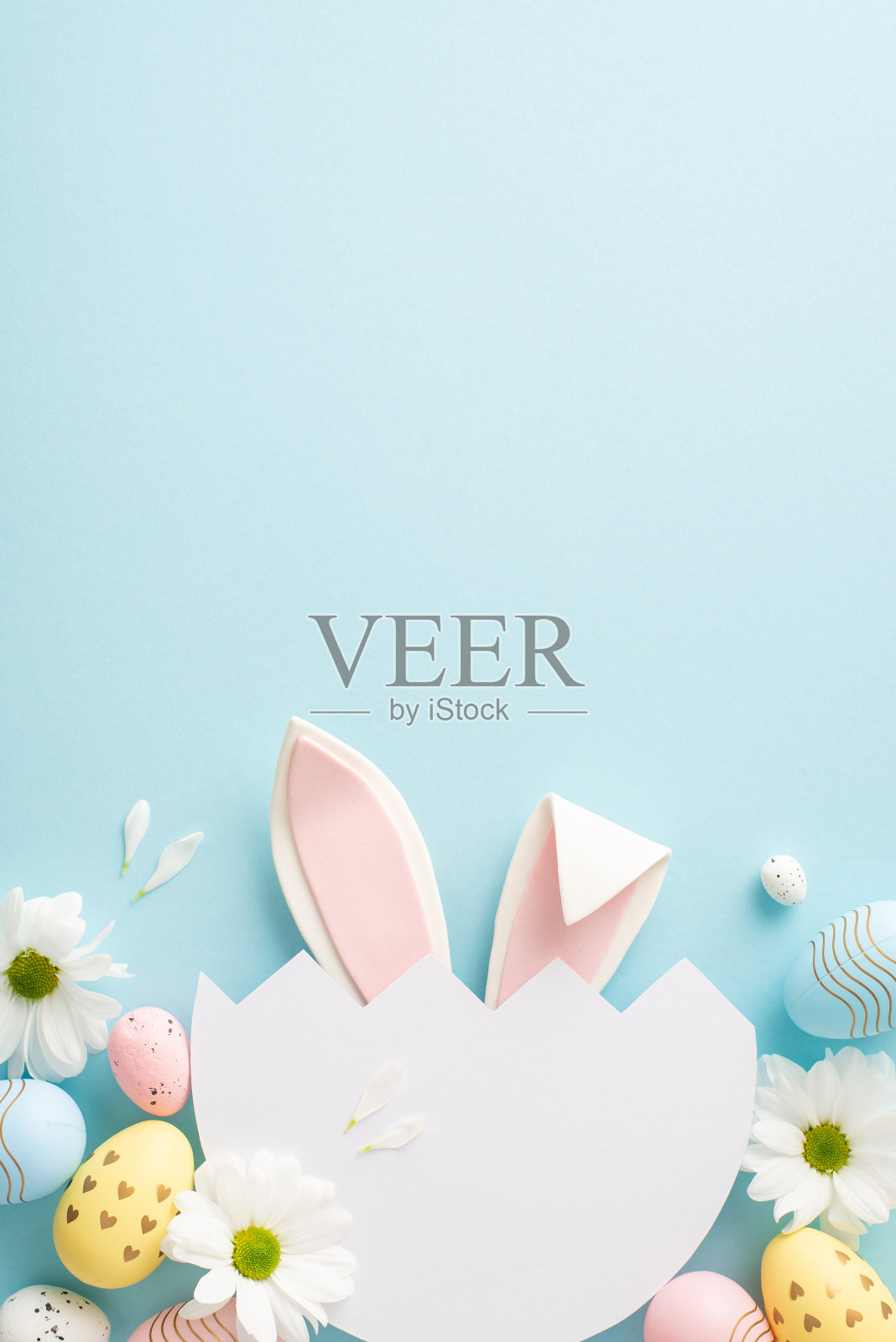 令人愉快的复活节惊喜概念。头顶上的垂直镜头，古怪的兔子耳朵从一个破鸡蛋里冒出来，周围是柔软的蓝色背景上的雏菊，留下空间给你的节日信息照片摄影图片
