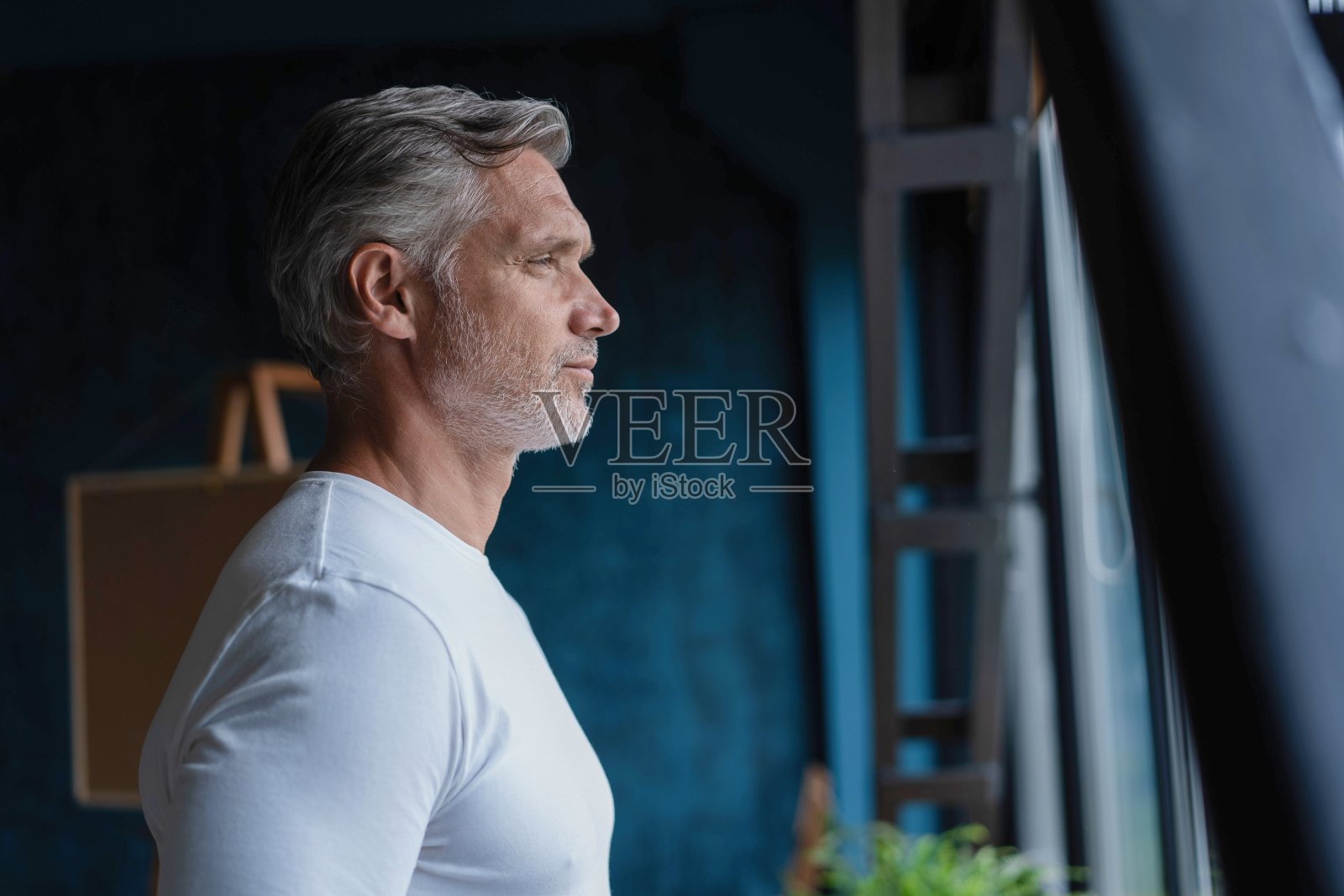 有魅力的成熟男人靠在一扇大而明亮的窗户的窗台上，看着别处。照片摄影图片