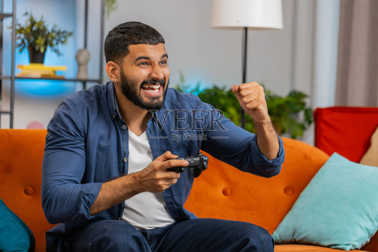 一名印度男子坐在家里的沙发上，用摇杆控制器玩视频游戏机，赢得了在线游戏的胜利照片摄影图片