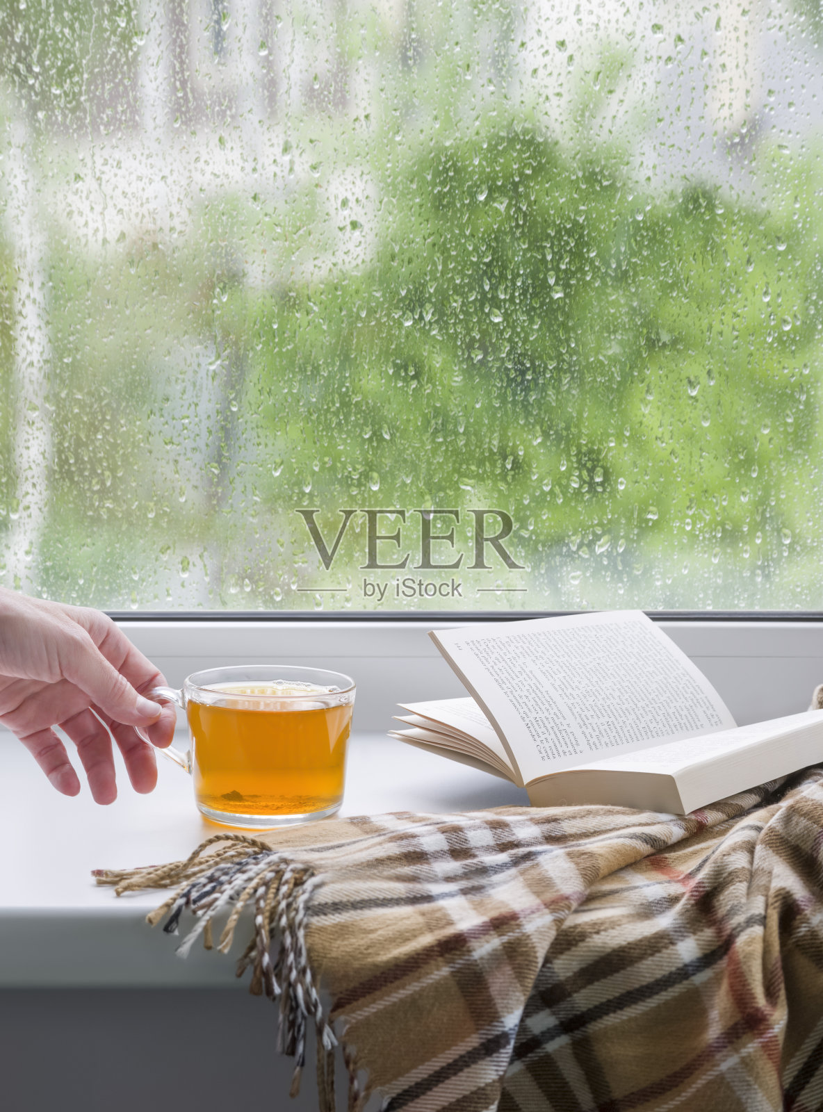 一个女人的手拿着一杯茶。一本打开的书和一条柔软的毯子。窗玻璃上布满了雨滴。春天照片摄影图片