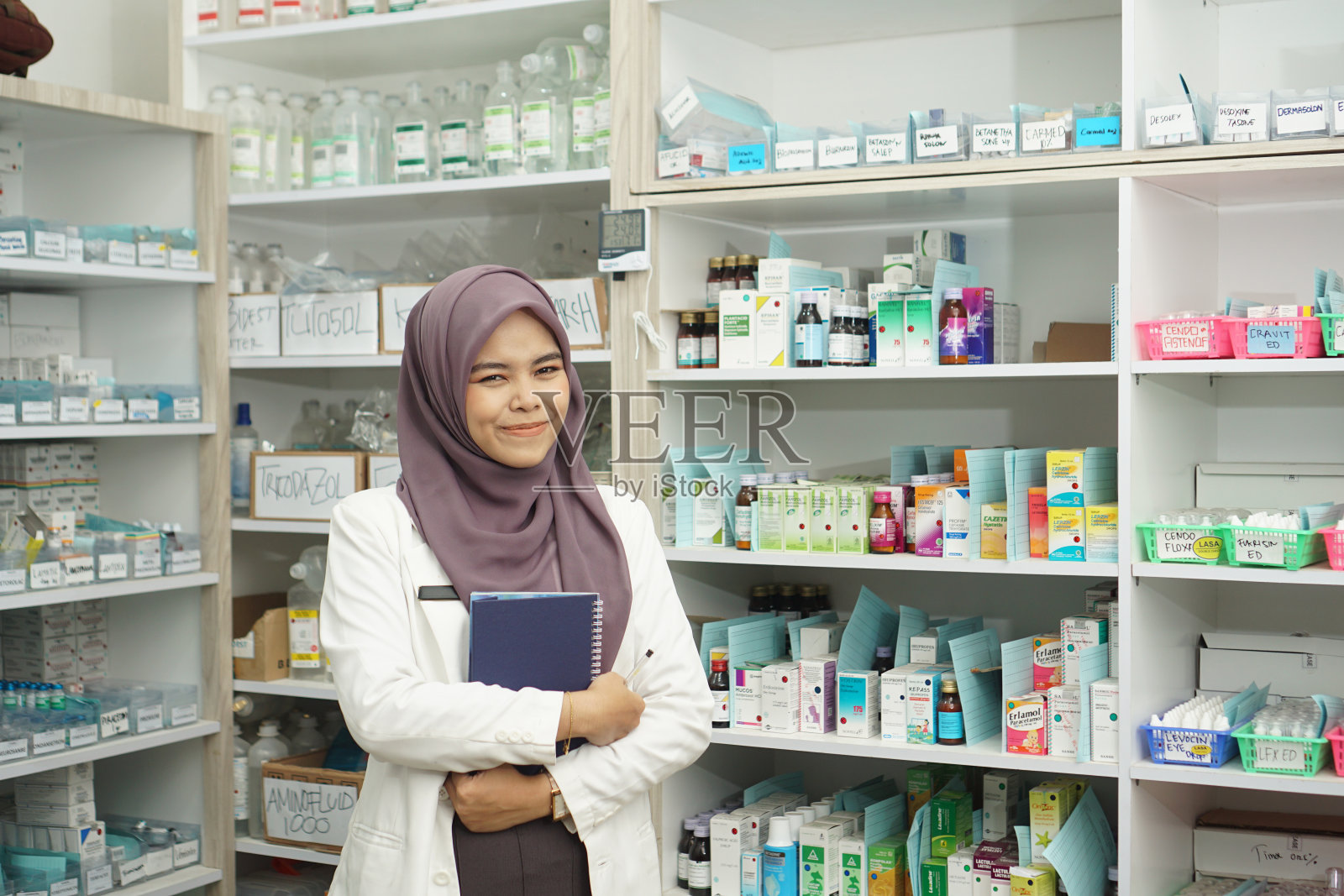 单身的印尼穆斯林女药剂师拿着书，微笑着站在药房里照片摄影图片