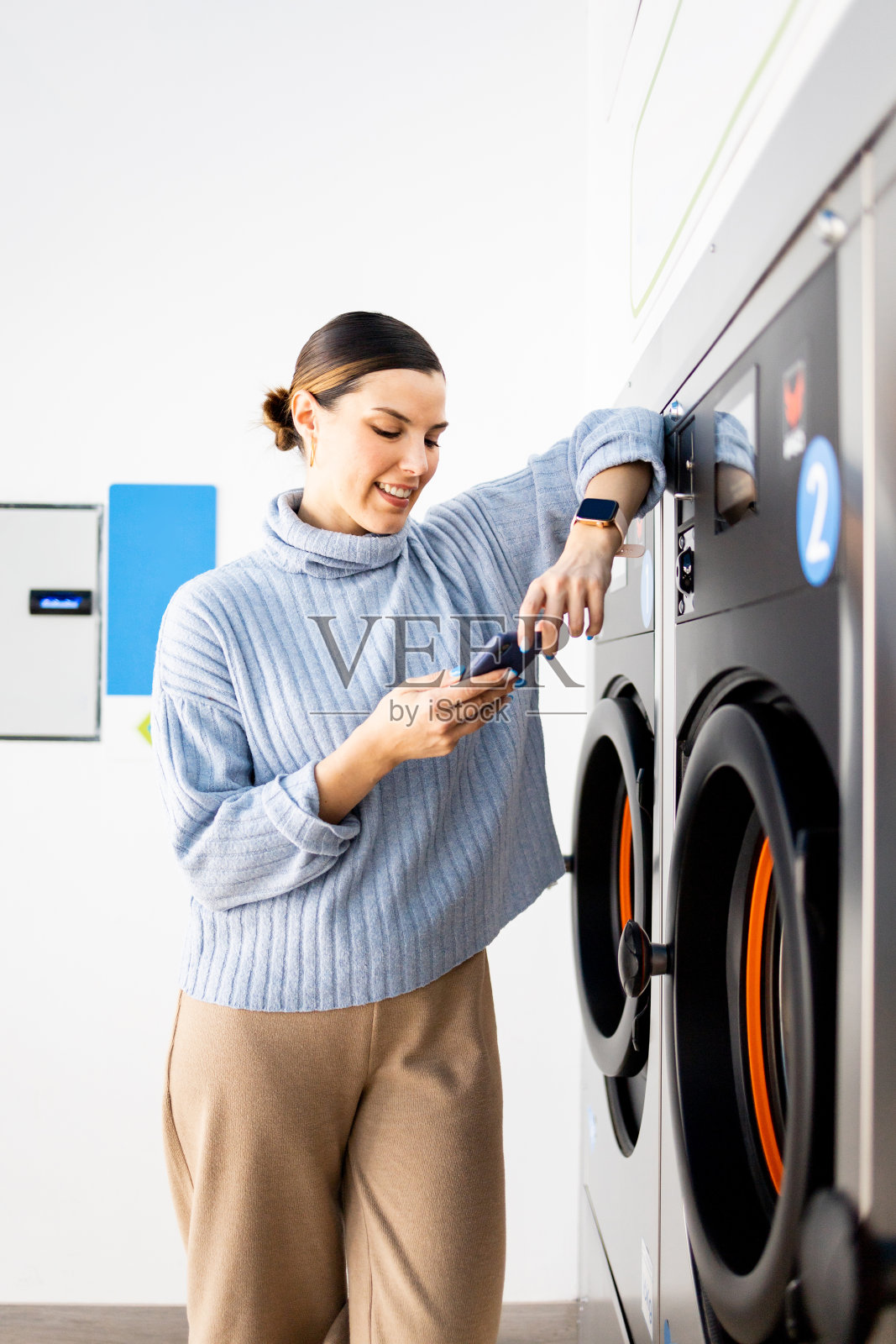 一个漂亮女孩站在自助洗衣店里的垂直照片。这名女子一边使用手机，一边靠在洗衣机上等待洗衣服。照片摄影图片