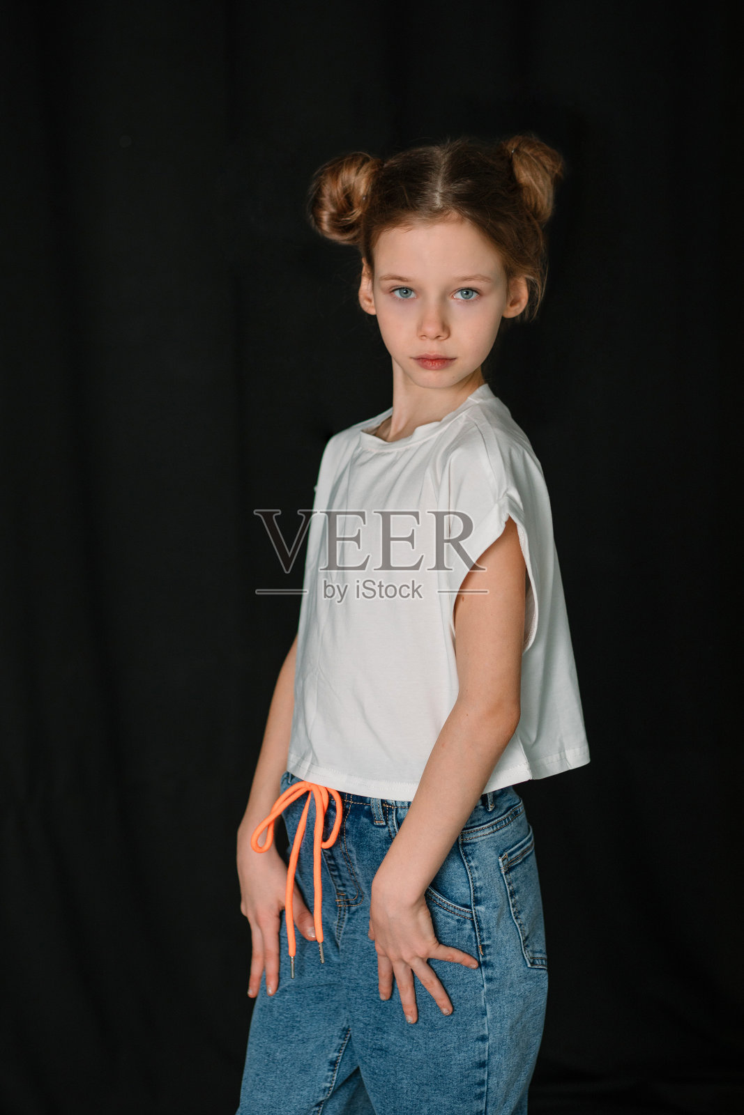 一个穿着白色t恤和蓝色牛仔裤的女孩站在黑色背景上。两个丸子头的女孩。儿童心理学。孩子和父母之间的关系。成人和青少年心理学。时髦的少女。恃强凌弱。照片摄影图片