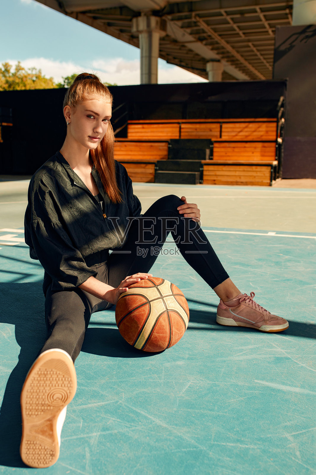 女篮球运动员微笑着坐在运动场上，拿着篮球看着镜头。运动、健身、积极的生活方式。照片摄影图片