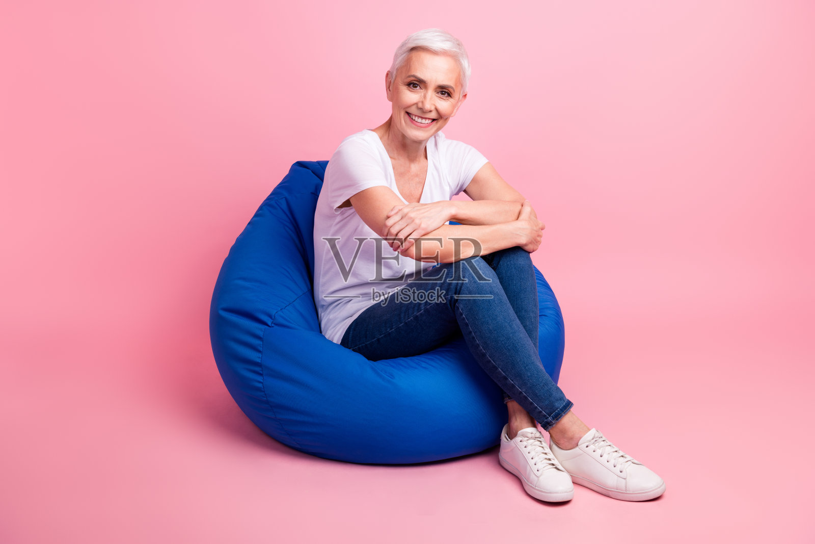 舒适的漂亮的退休妇女穿白色t恤牛仔裤坐在舒适的豆袋孤立在粉红色的背景照片摄影图片