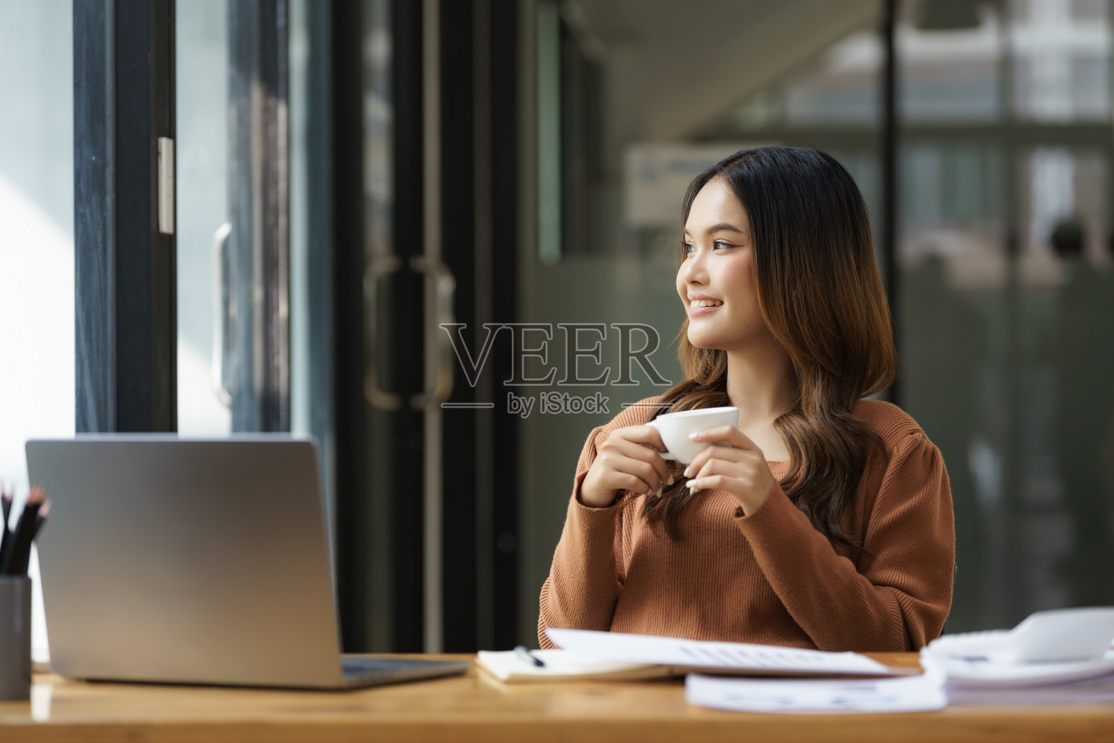 一位年轻的亚洲女商人坐在办公室里，端着一杯白色的咖啡。照片摄影图片