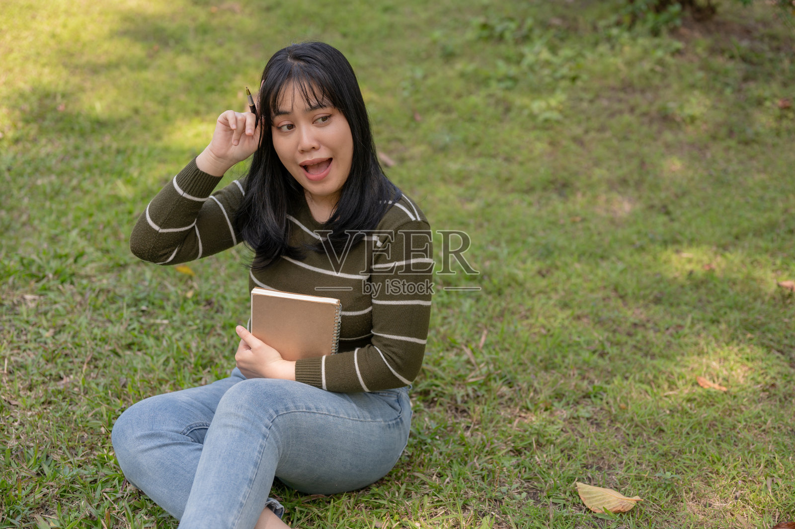一个积极向上的年轻亚洲女人坐在公园的草地上，拿着笔记本，思考着新的想法。照片摄影图片