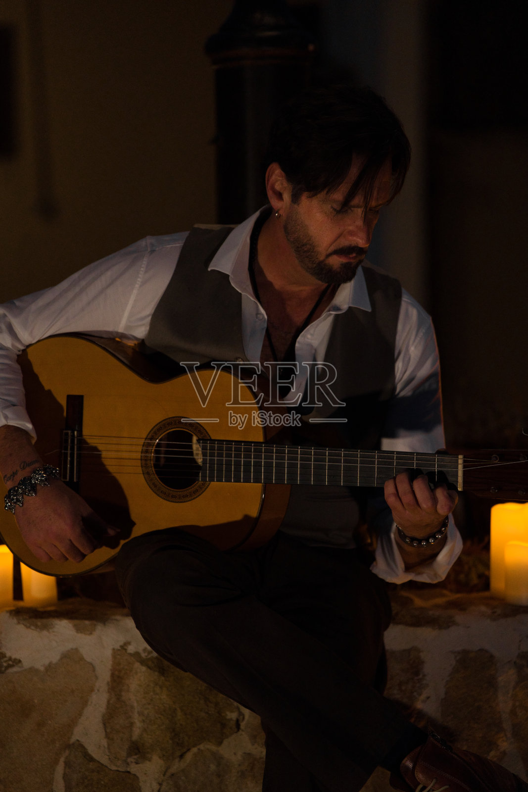 一个穿着黑白马甲的专业吉他手在夜晚的村庄广场上举行烛光音乐会的垂直照片。照片摄影图片