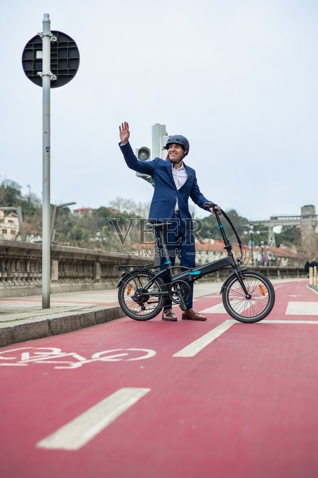 身穿蓝色西装、头戴头盔的商人站在红色的自行车道上，手里拿着电动折叠自行车，向远处挥手照片摄影图片