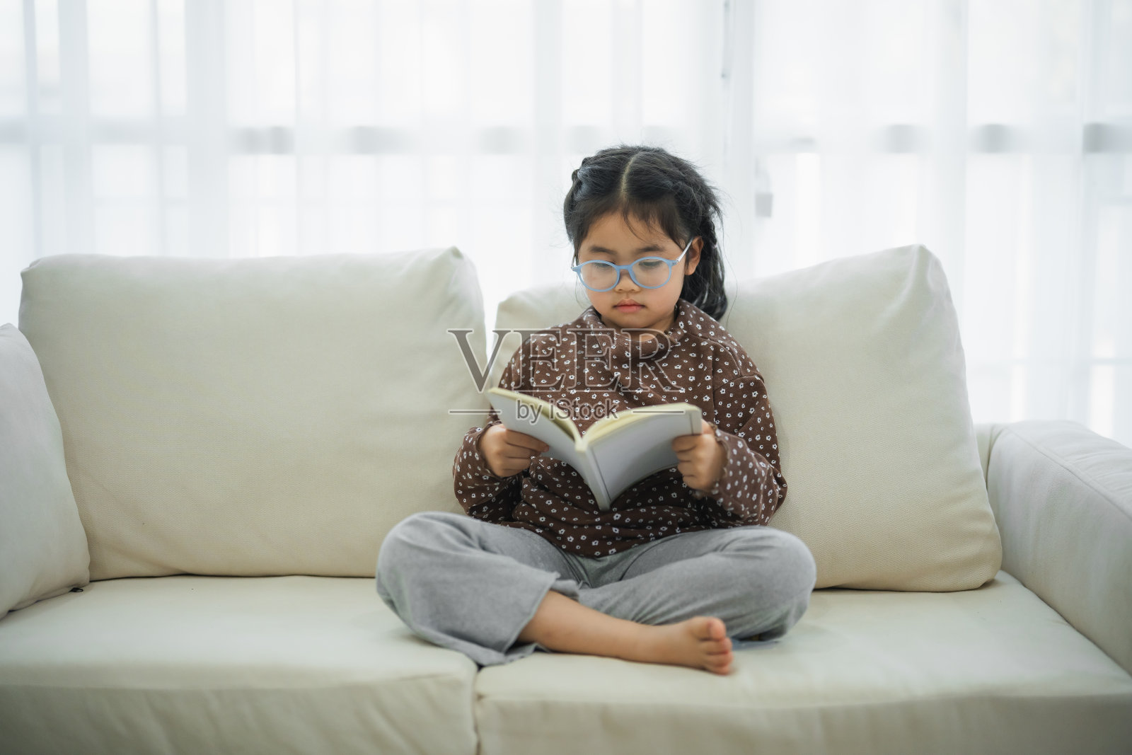 快乐的亚洲孩子，小女孩微笑着戴着眼镜，坐在家里客厅的沙发上看书。女孩放松看书，在家里的沙发上微笑。回到学校的概念。照片摄影图片