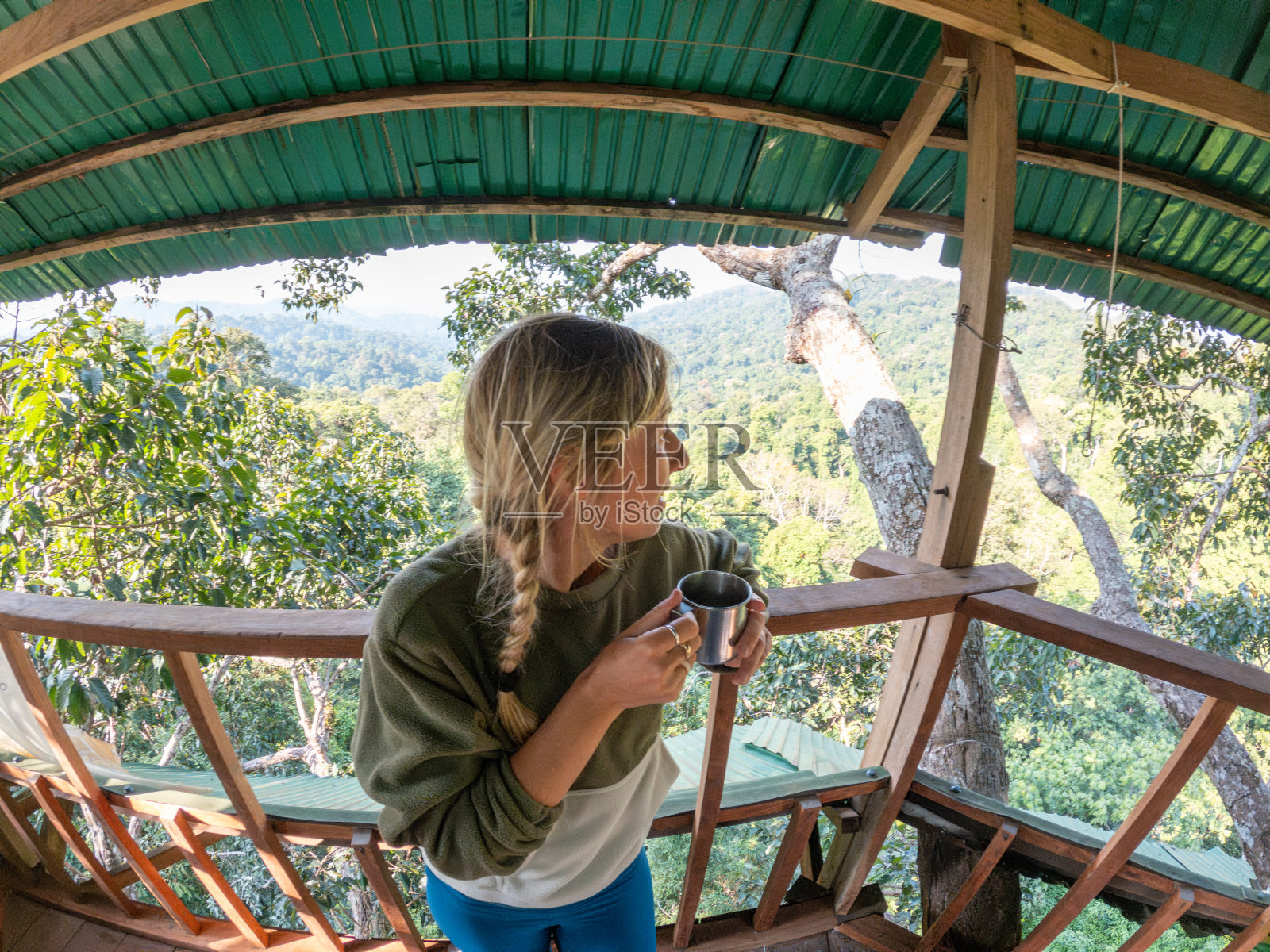 高空反射:一个年轻的女人从树屋里俯瞰丛林，手里拿着旅行马克杯，看着下面的大自然照片摄影图片