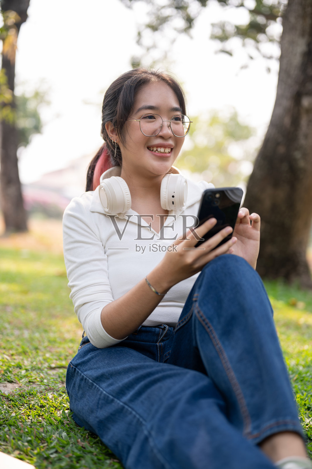 一个美丽快乐的亚洲女人坐在绿色公园的草地上玩着她的智能手机。照片摄影图片