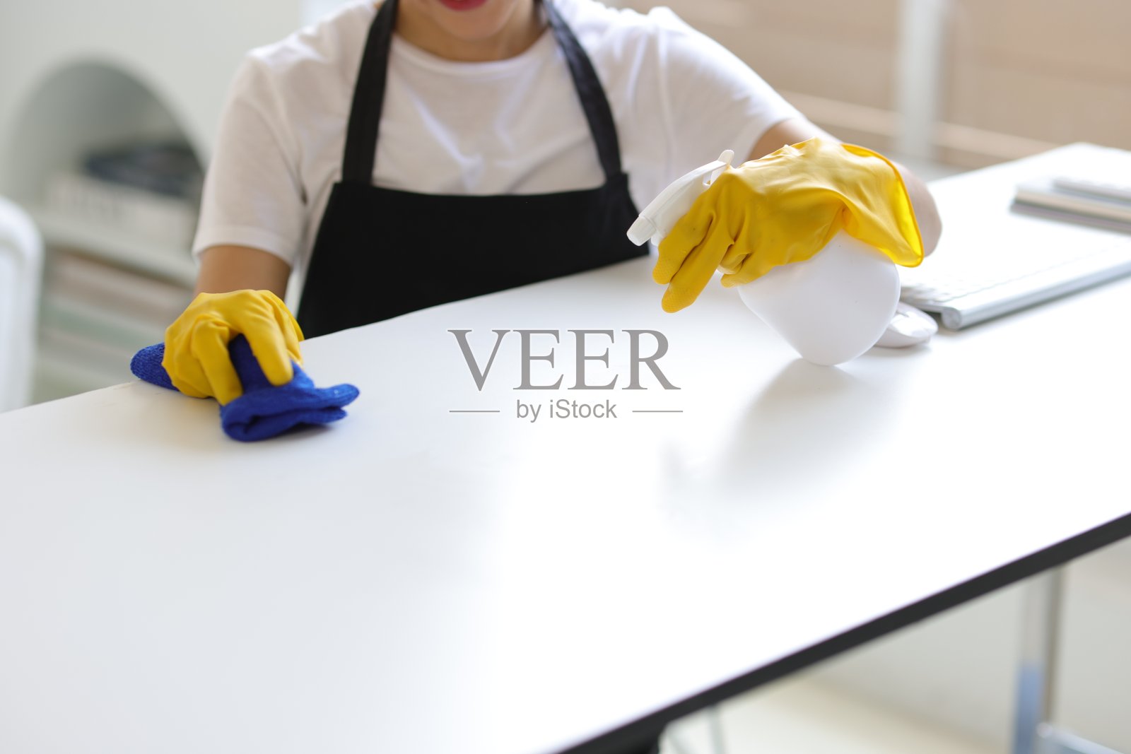 一位系着围裙的年轻女子正在家庭办公室用消毒液和抹布擦拭桌子。照片摄影图片