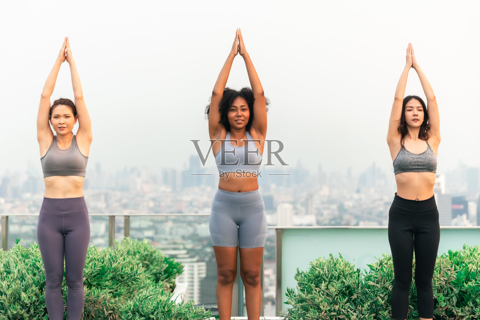 一群年轻人在室内瑜伽课时冥想观想。年轻的亚洲妇女和非裔美国人在屋顶上做瑜伽。一群快乐健康的朋友在城市户外锻炼。照片摄影图片