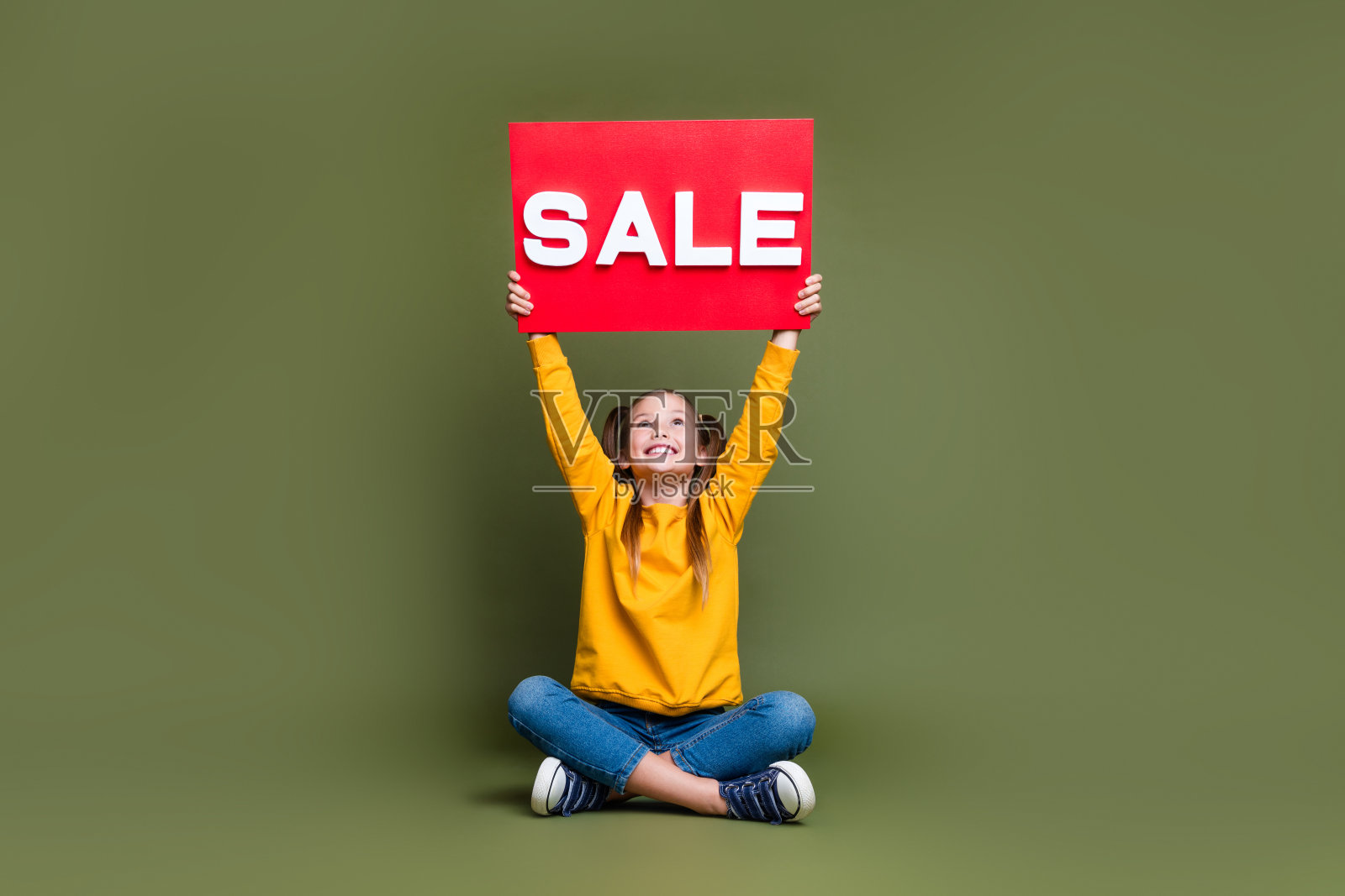 漂亮的女学生的全尺寸照片与尾巴穿黄色套头衫看销售板在头上孤立的卡其色背景照片摄影图片