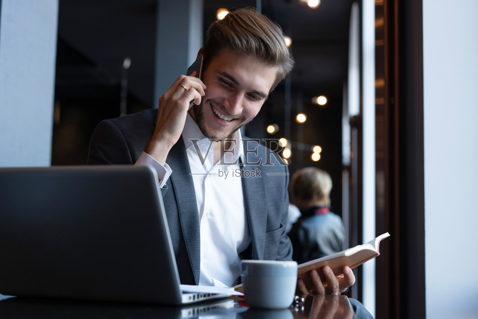 开朗的商人穿着正装坐在咖啡馆里，一边打电话一边用笔记本电脑。照片摄影图片