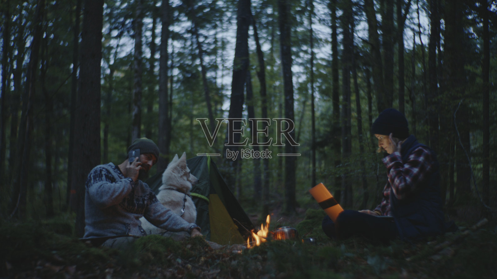 在森林篝火旁，男人在和狗通电话，女人在用笔记本电脑工作照片摄影图片