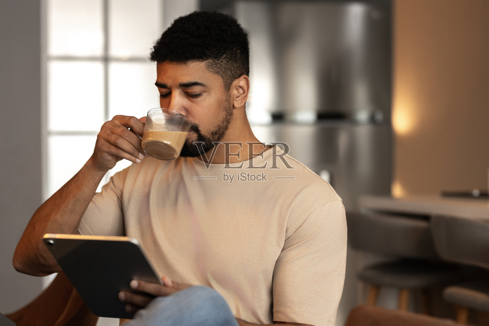 英俊的年轻黑人，穿着休闲装，坐在客厅的椅子上，一边喝咖啡，一边用平板电脑查看社交网络照片摄影图片