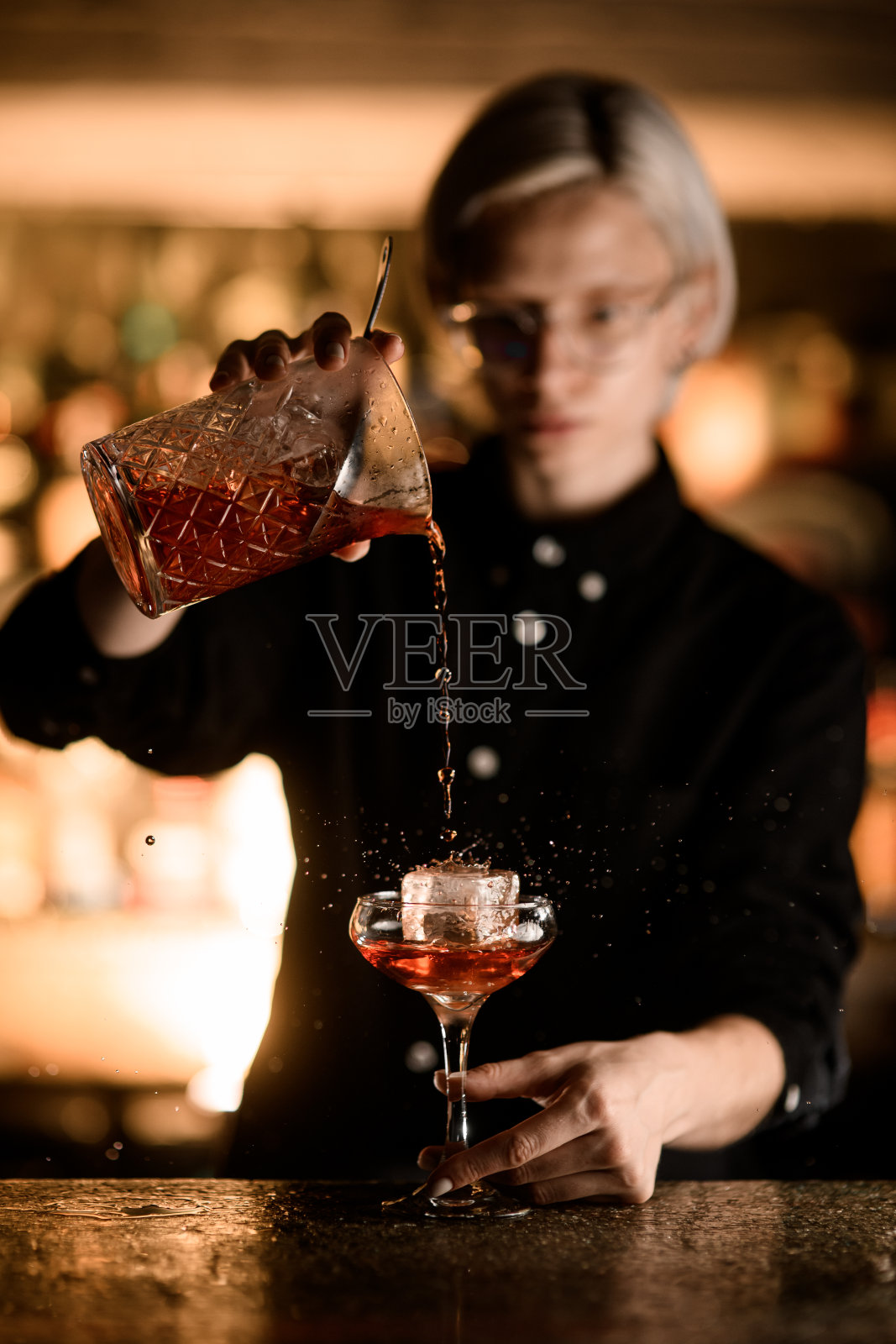 女调酒师将一杯鸡尾酒倒入加了冰的玻璃杯中，这样饮料就会溅到四周照片摄影图片