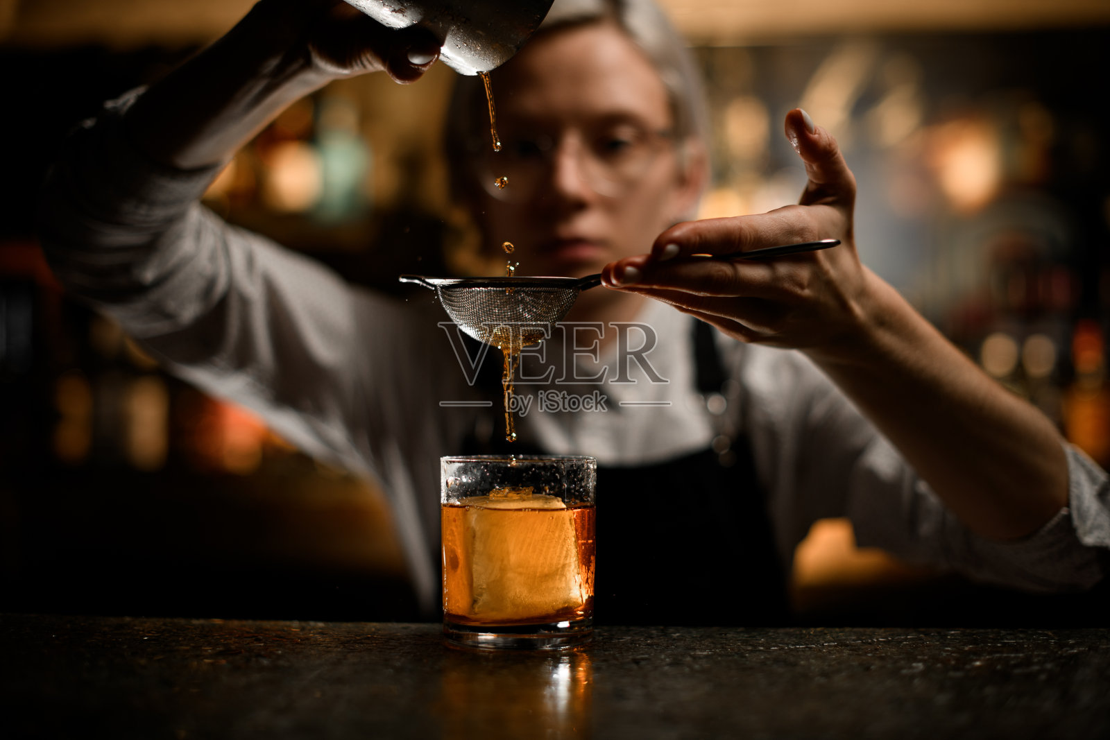 女调酒师把最后几滴鸡尾酒从一个金属玻璃杯里倒进加了冰的玻璃杯里照片摄影图片