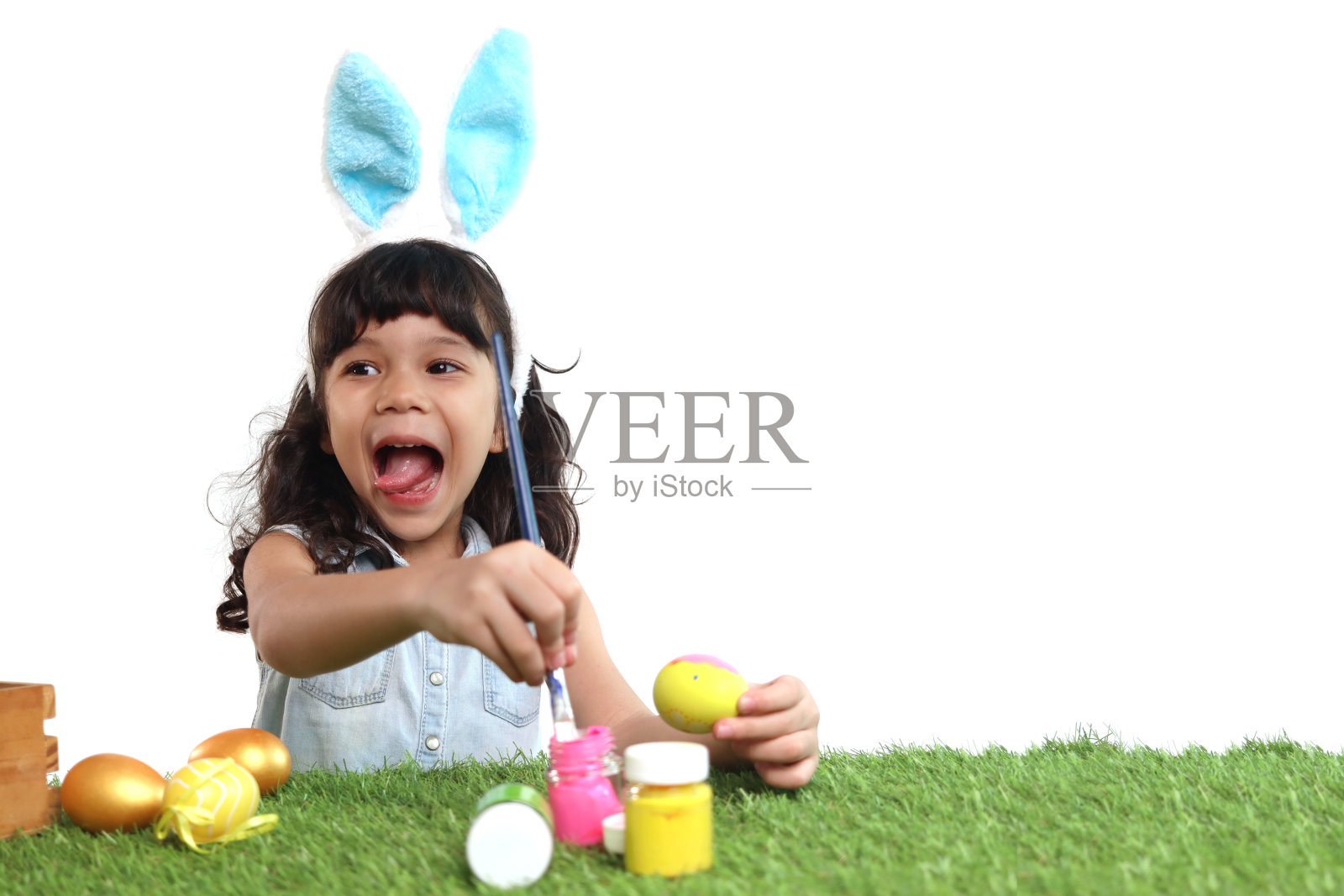 可爱的小女孩长着蓝色的兔子耳朵，用刷子在绿色的草地上画鸡蛋，快乐的孩子在白色的背景上庆祝复活节。快乐的孩子在复活节庆祝活动中玩得很开心。照片摄影图片