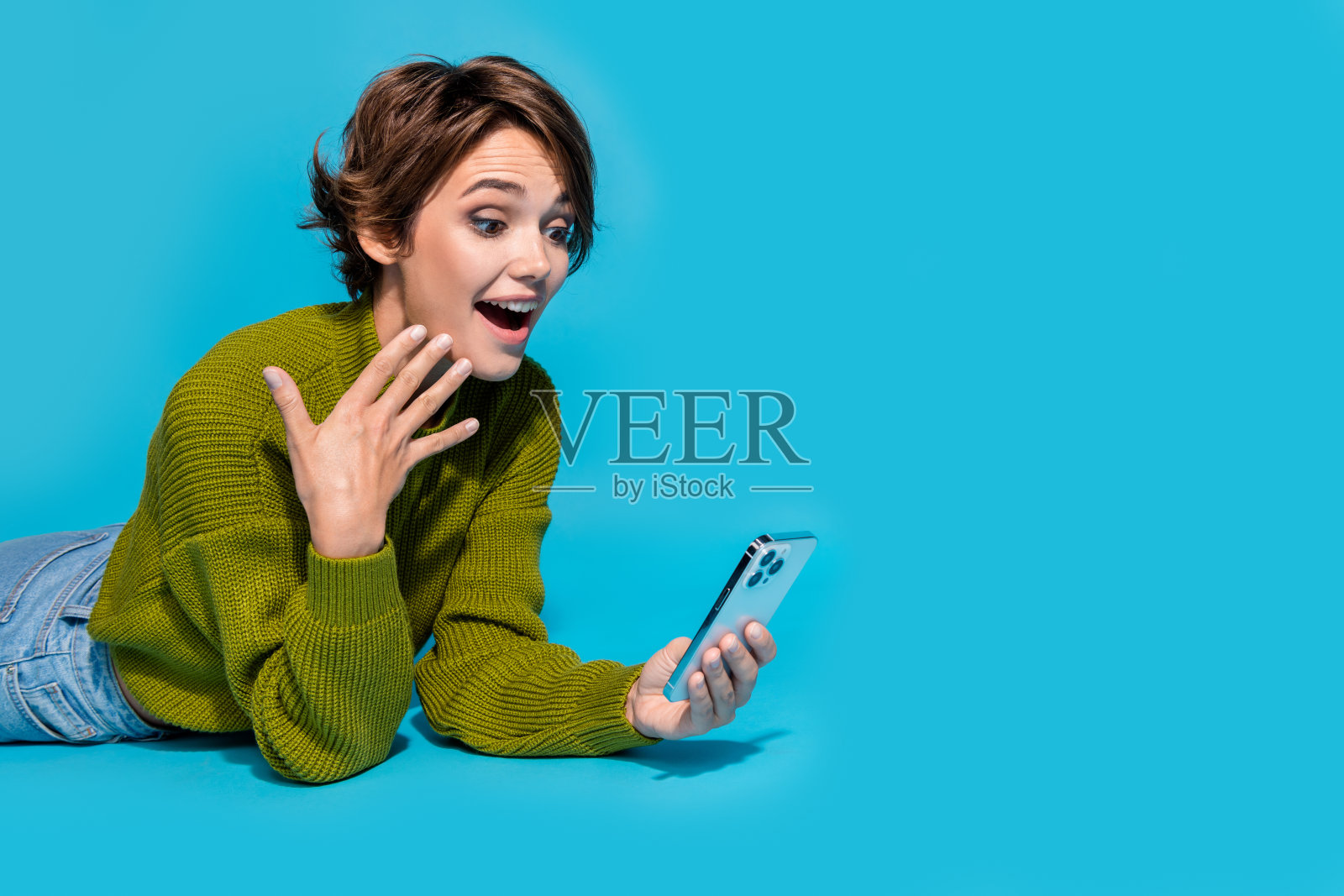 照片中迷人、惊讶、震惊的女孩穿着时髦的衣服躺在地板上聊天，在青色的背景下孤立的空间照片摄影图片