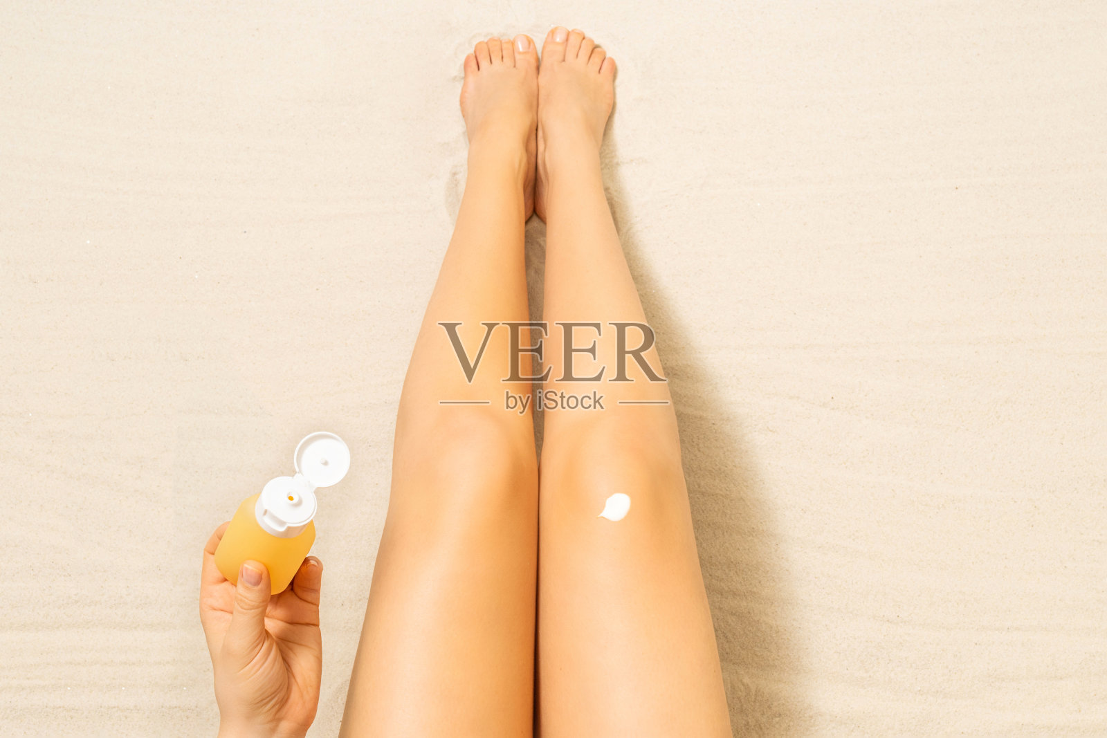 沙滩上苗条的女人腿的俯视图。女性坐在沙滩上晒日光浴，涂防晒霜保护皮肤。暑假放松。鸟瞰图。照片摄影图片