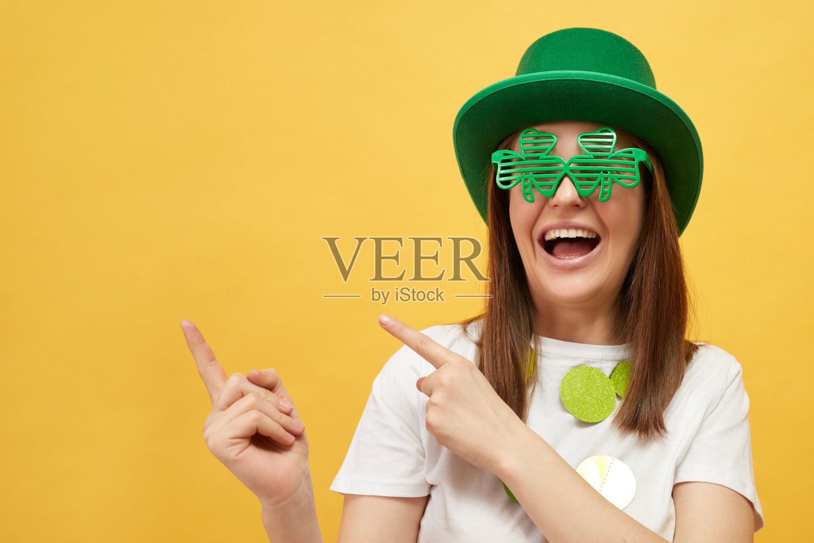 有趣的圣帕特里克节庆祝活动。戴着绿色小妖精帽子和三叶草眼镜的女人微笑着，喜出望外，在黄色的背景上孤立地摆姿势，指着广告的复制空间照片摄影图片