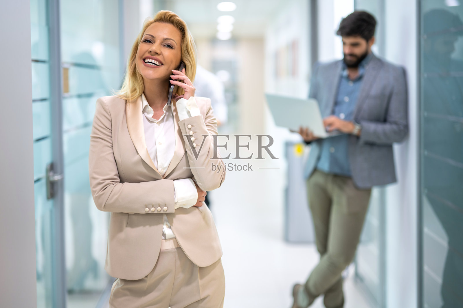 年轻的金发女商人在办公室走廊上讲手机的画像。照片摄影图片