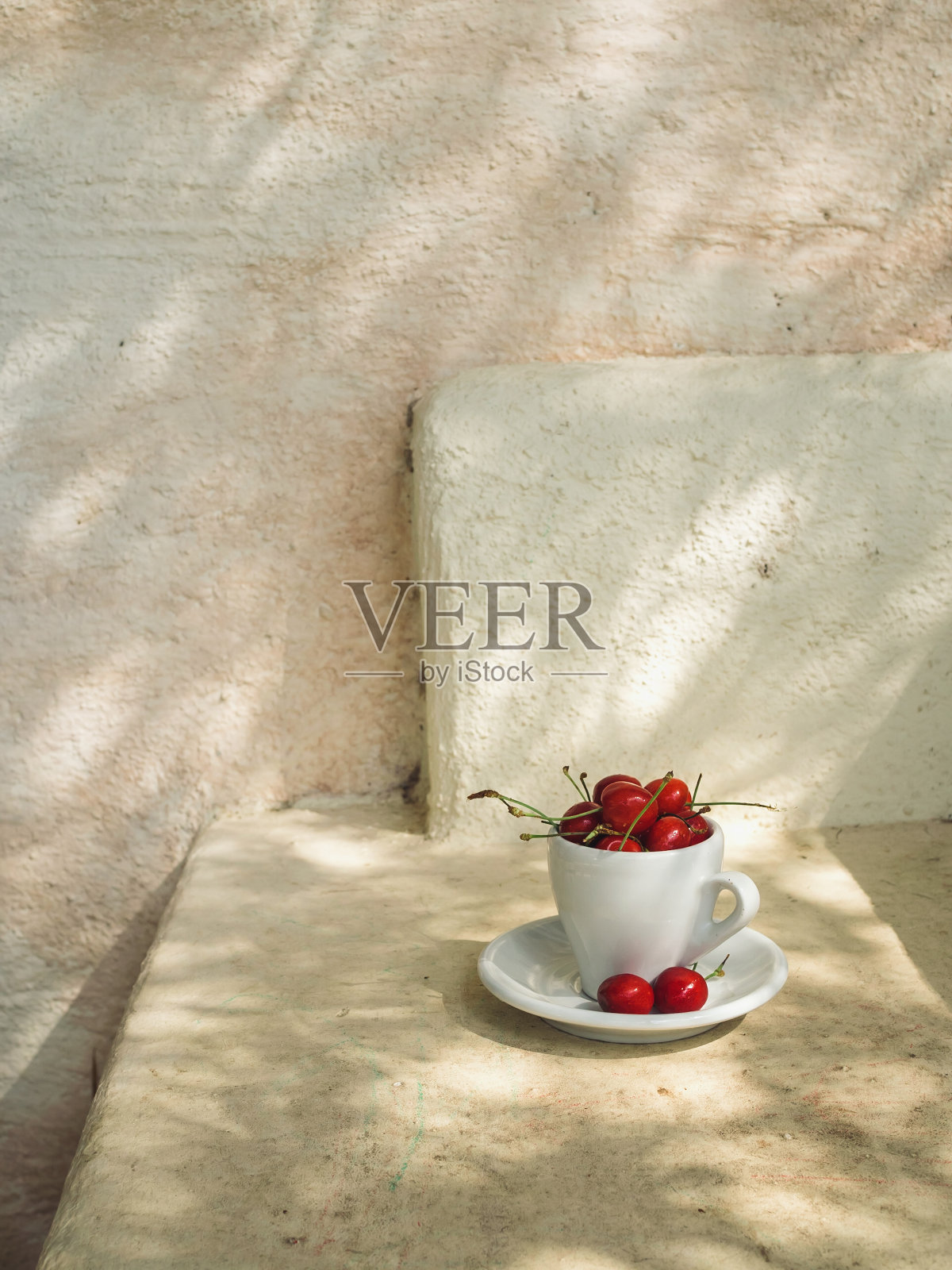 垂直特写镜头的樱桃在一个白色的咖啡杯和茶碟上的米色意大利阳台表面在自然模式的橄榄树的阴影。照片摄影图片