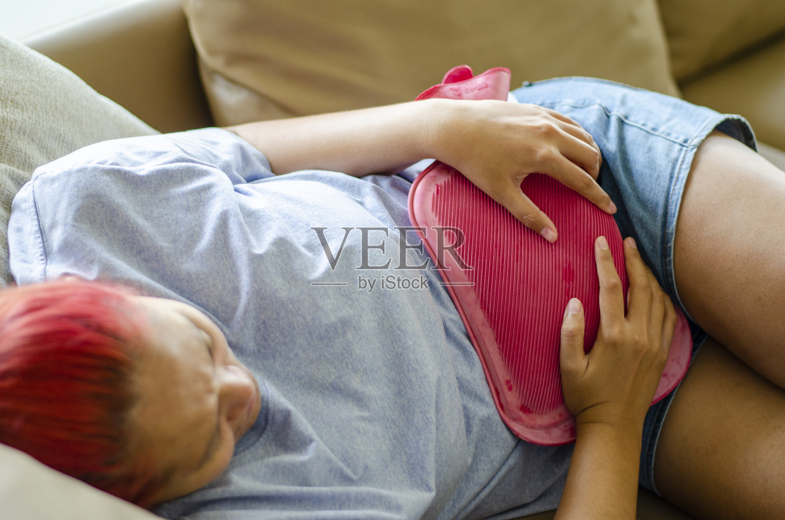 年轻的亚洲女子躺在沙发上，用热水袋来缓解痛经。照片摄影图片