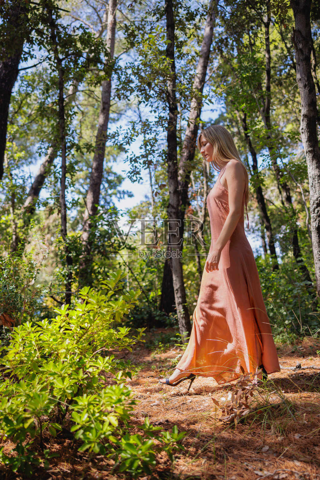漂亮的金发女人穿着赤陶色无袖长裙独自在树林里游荡照片摄影图片