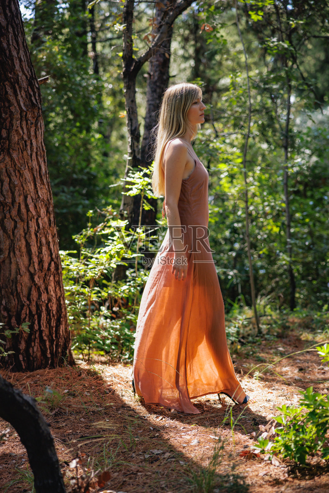 穿着赤陶色无袖连衣裙的金发美女独自漫步在阴凉的森林里照片摄影图片