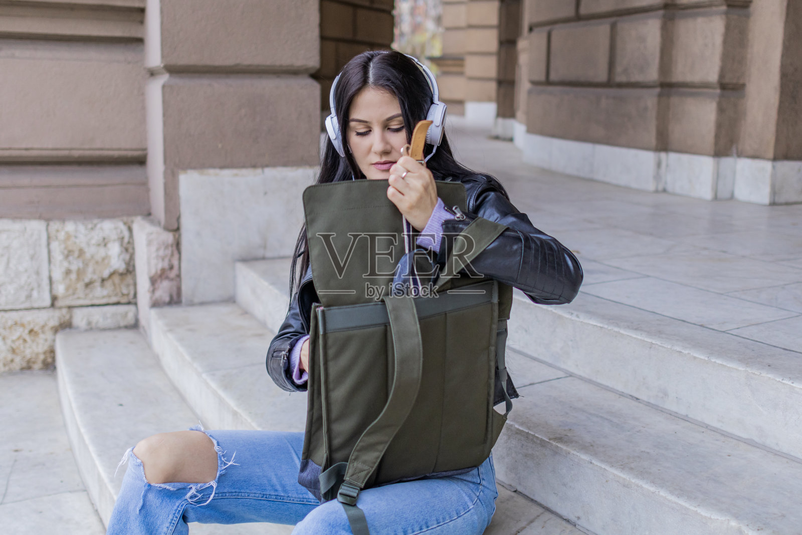 年轻漂亮的女大学生背着双肩包坐在大学楼梯上。她戴着耳机，穿着皮夹克，等待着大学课程的开始。照片摄影图片